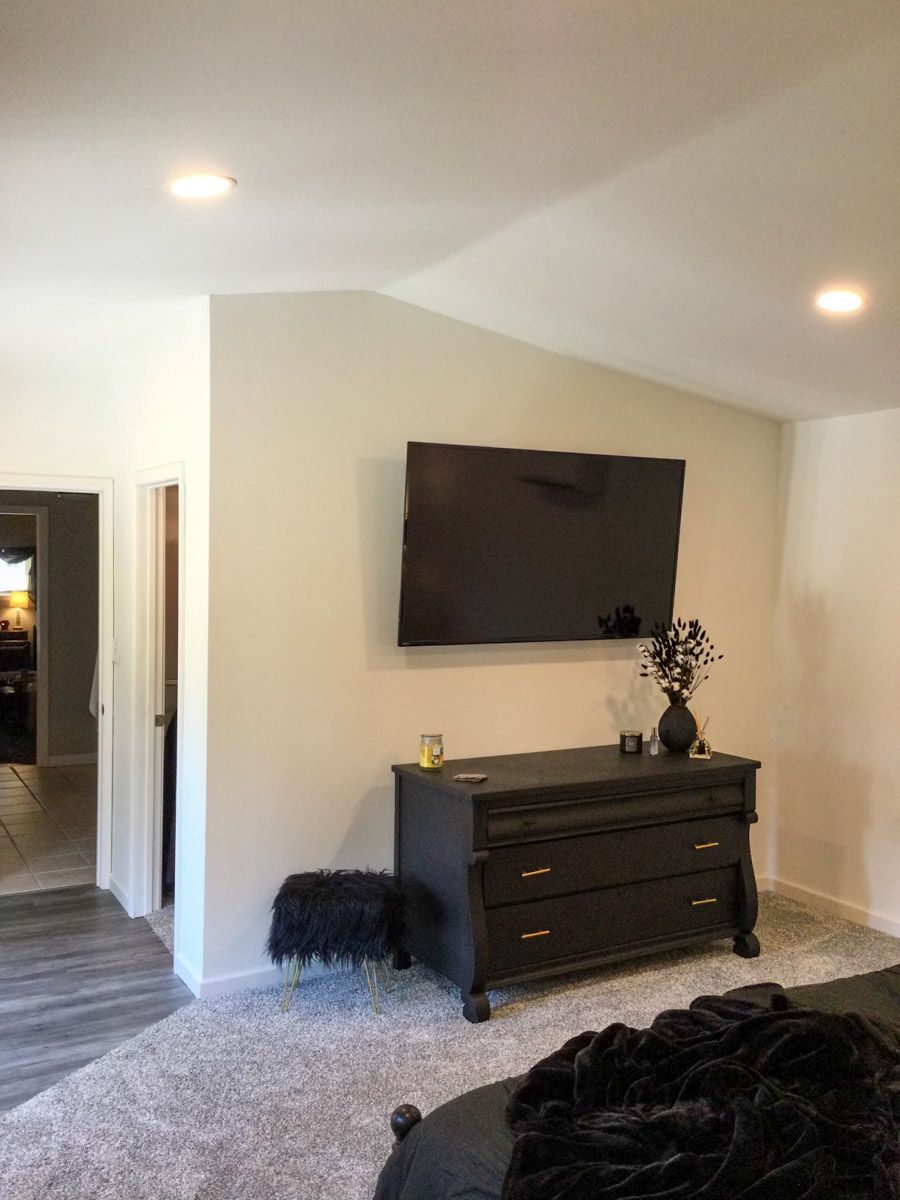 Black TV mounted above a black dresser with gold handles in a bedroom with gray carpet.