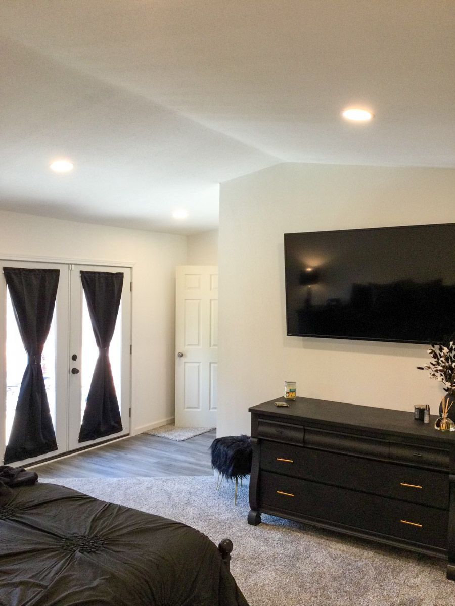 Bedroom with a black dresser, large TV, French doors with black curtains, and a bed.