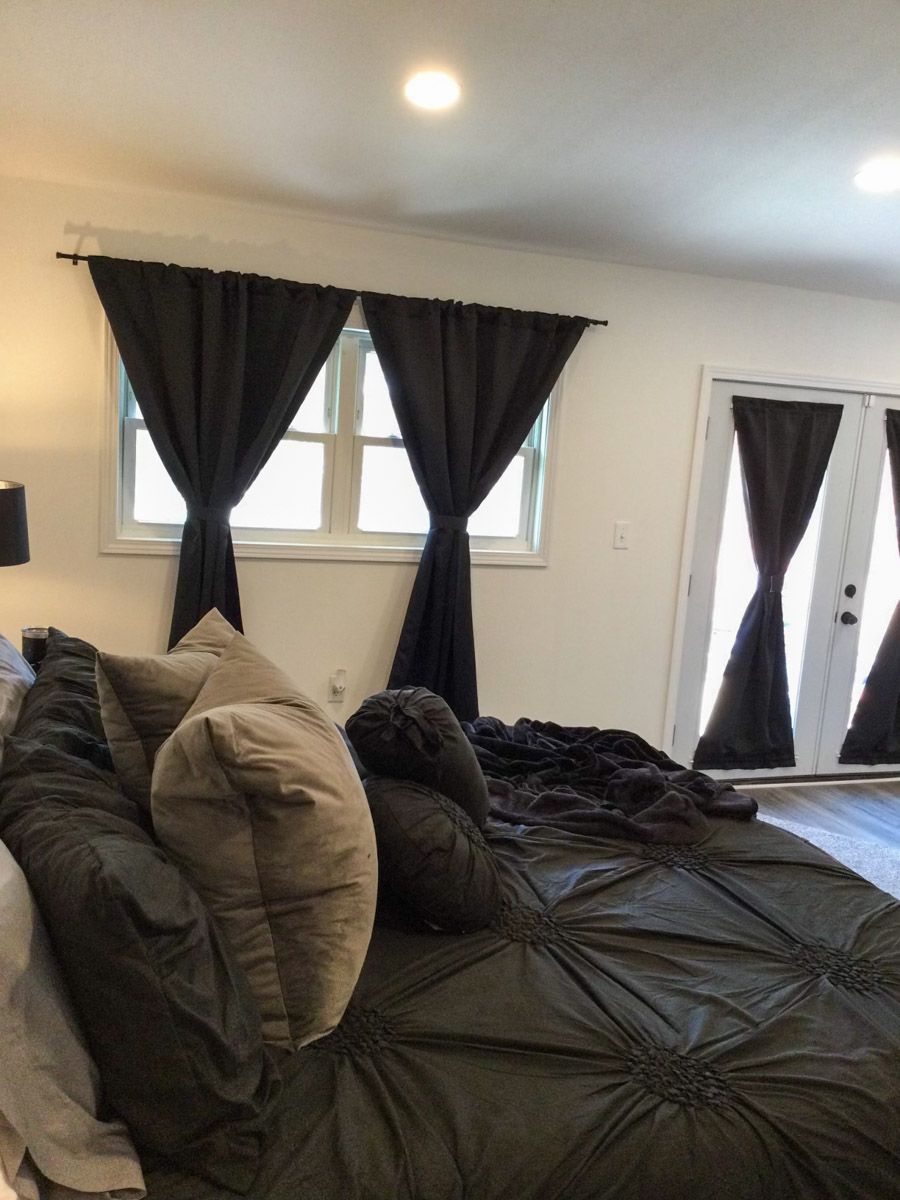 Dark bedroom with black curtains, white walls, and a large bed with pillows.