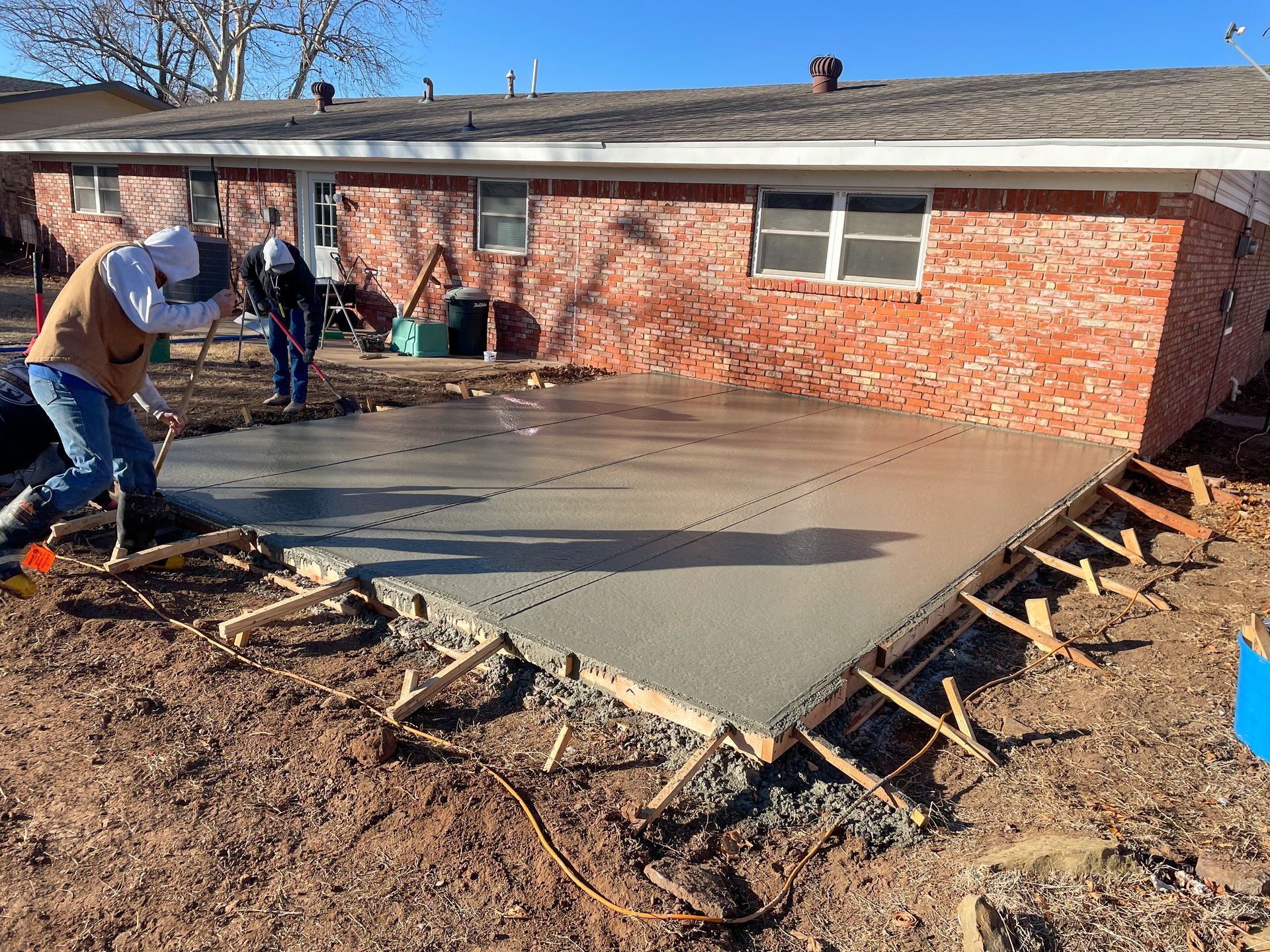 Workers pouring concrete patio next to a red brick house; wooden frame visible.