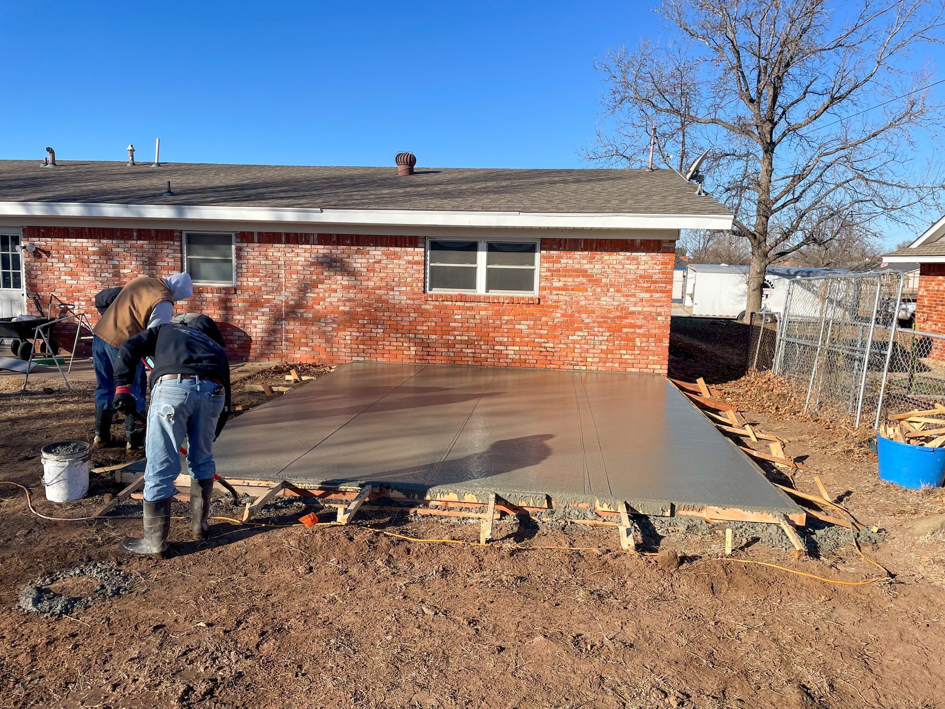 Workers pouring and smoothing concrete for a patio next to a red brick house; blue sky.