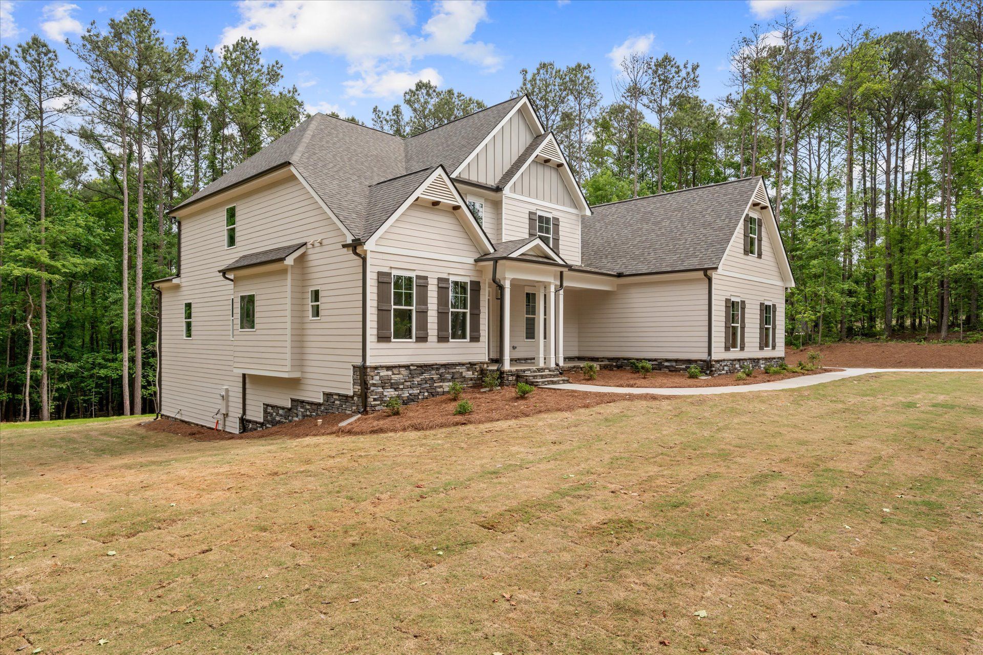 Beige suburban house with gabled roof, stone accents, and a wooded yard on a sunny day