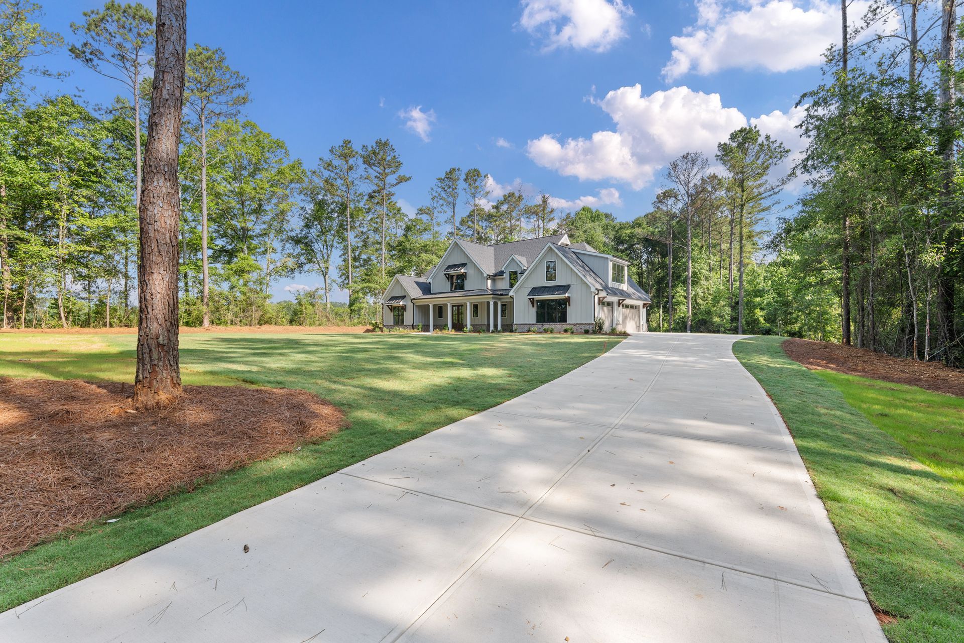 Long driveway leading to a gray house with a green lawn and tall pine trees under a blue sky