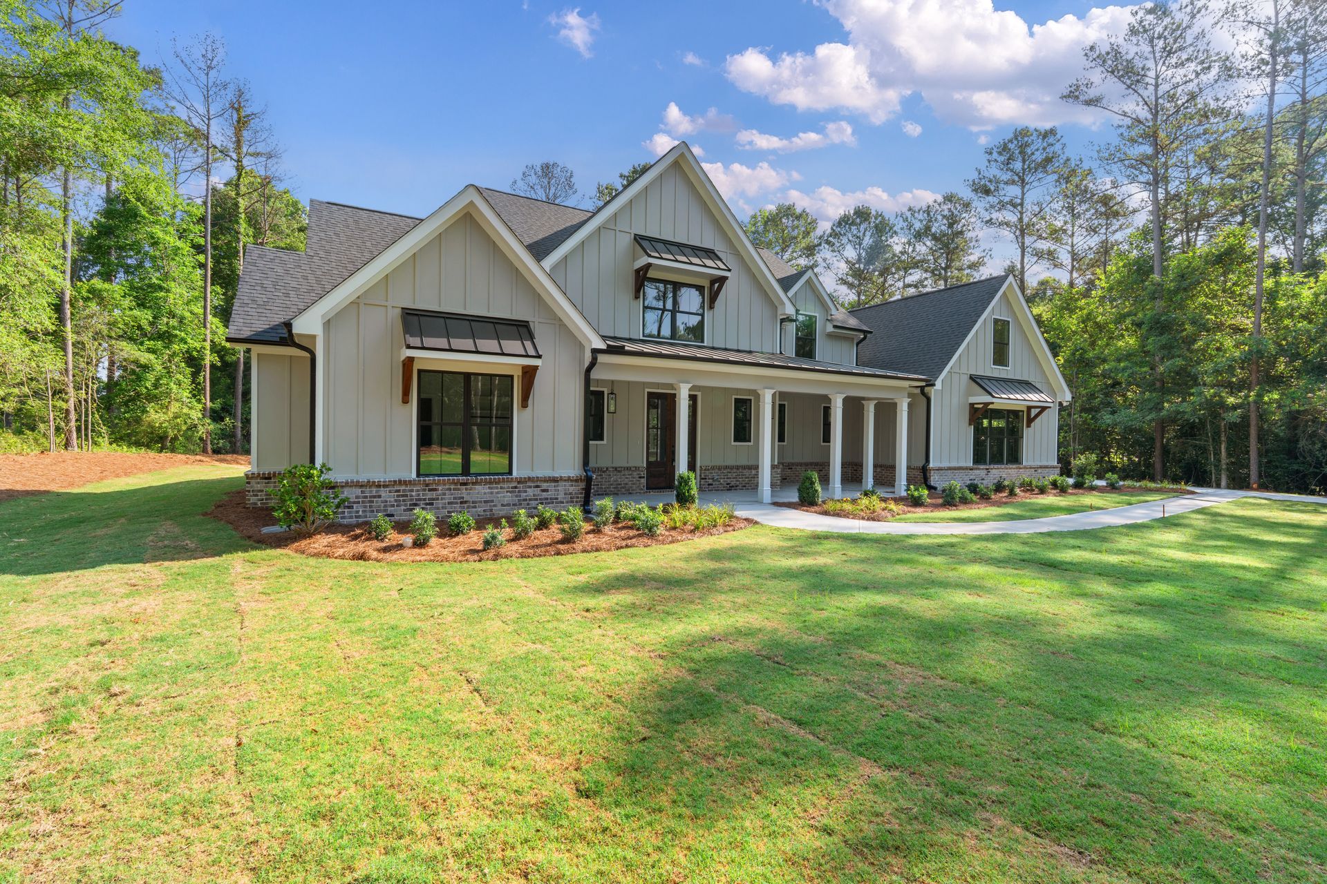 Large modern suburban house with gray roof, beige exterior, and a manicured front lawn under a blue sky