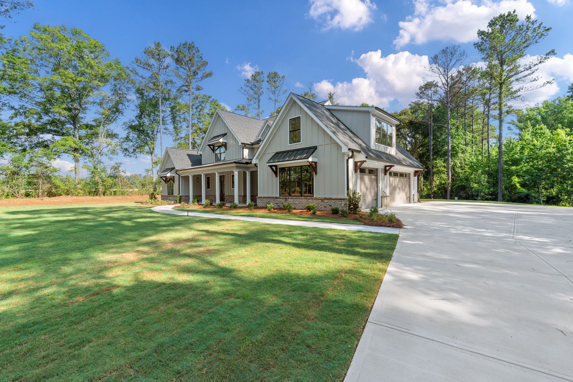 Large gray suburban house with a wide lawn, driveway, and trees under a blue sky