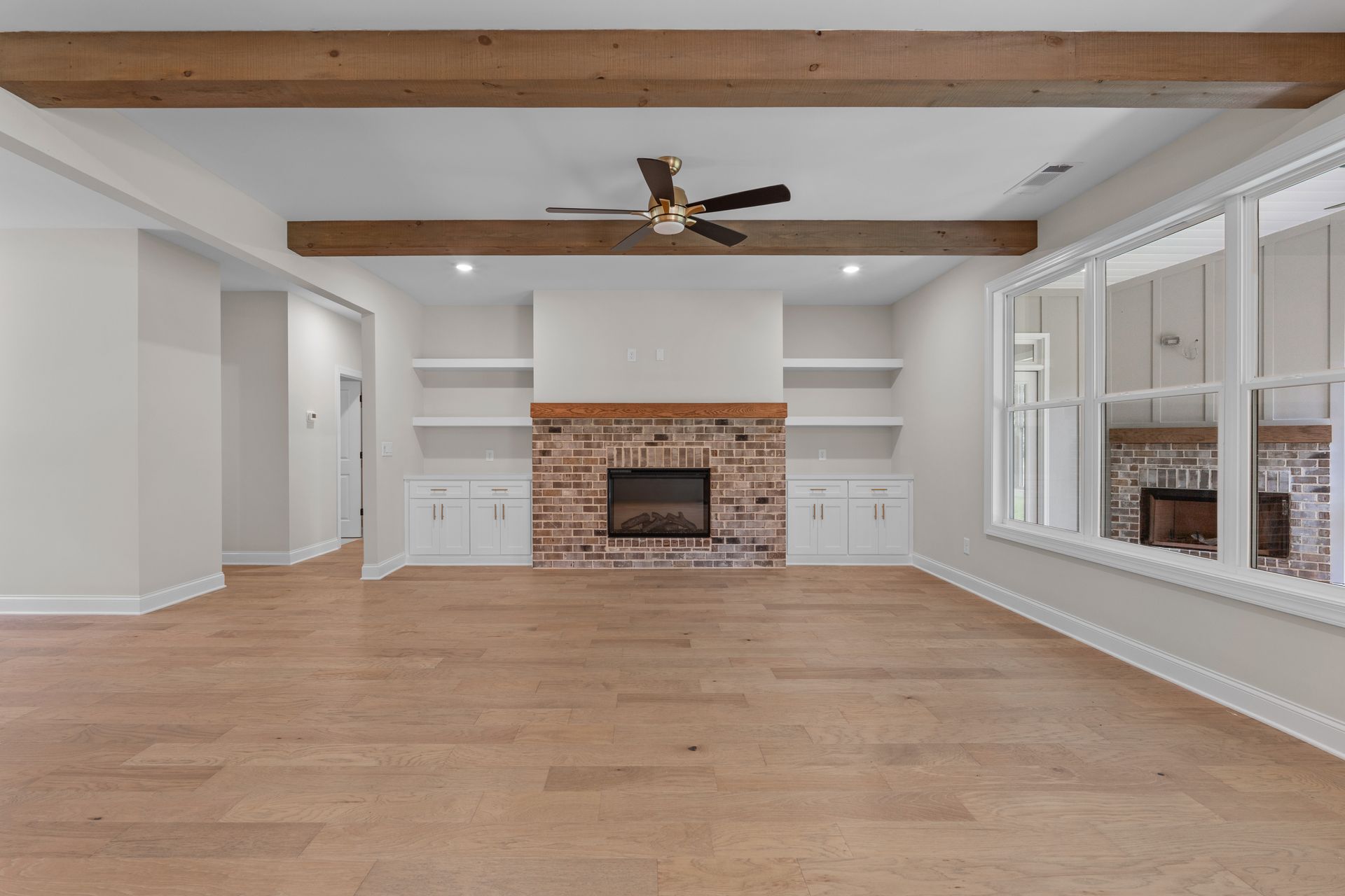Empty living room with hardwood floors, exposed beams, brick fireplace, built-in shelves, and large windows