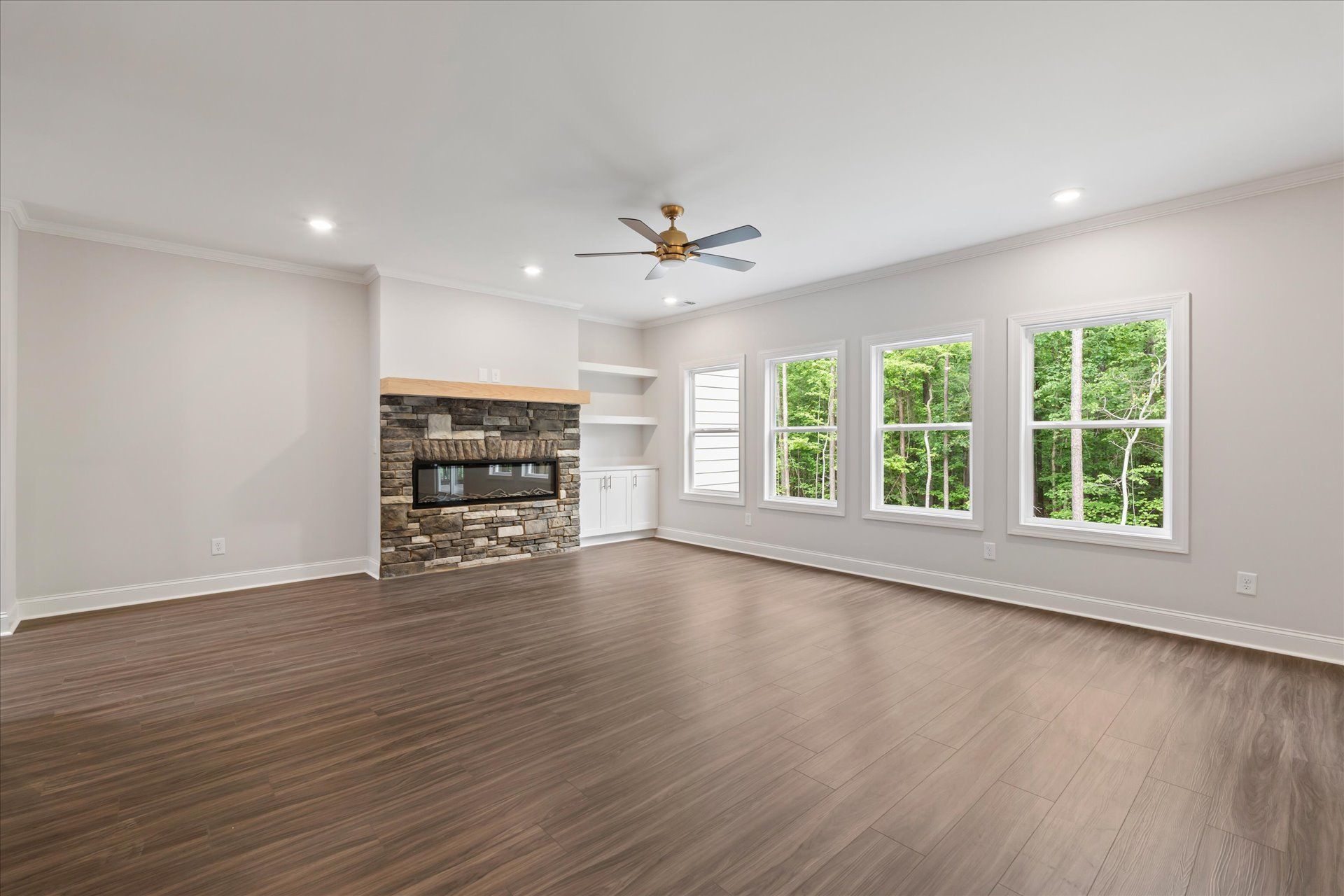 Spacious empty living room with wood floors, white walls, fireplace, and large windows overlooking greenery
