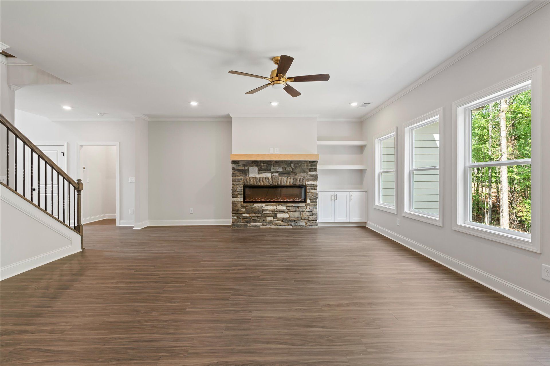 Spacious empty living room with wood floors, stone fireplace, ceiling fan, and large windows
