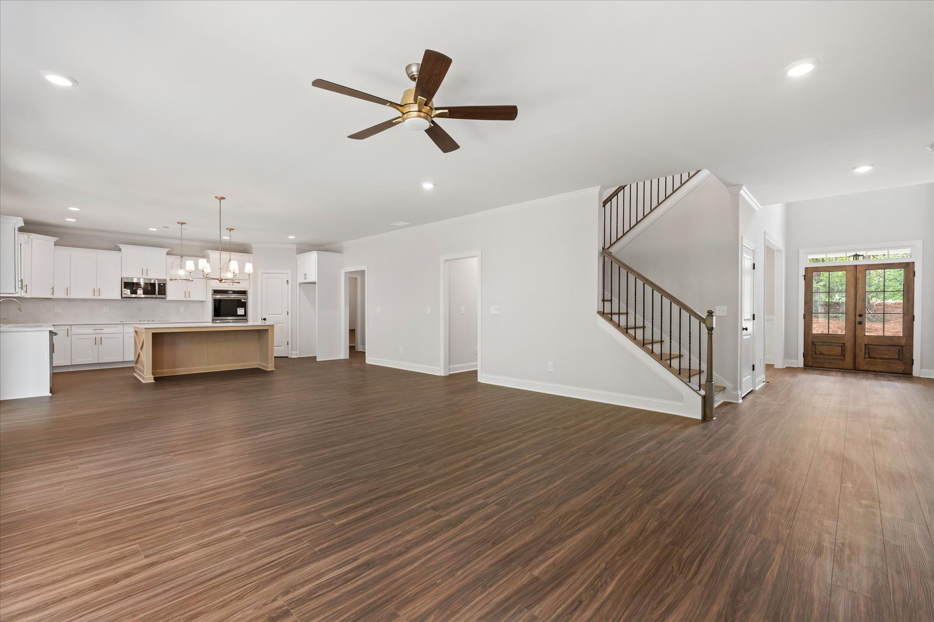 Empty open-plan living room with kitchen, wood floors, ceiling fan, and staircase by the entryway