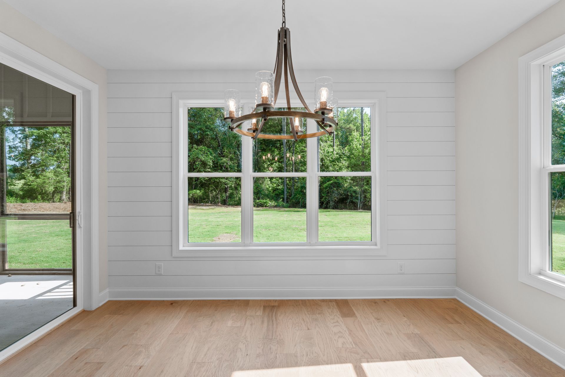 Bright empty room with white paneled walls, hardwood floor, and a chandelier under large windows.