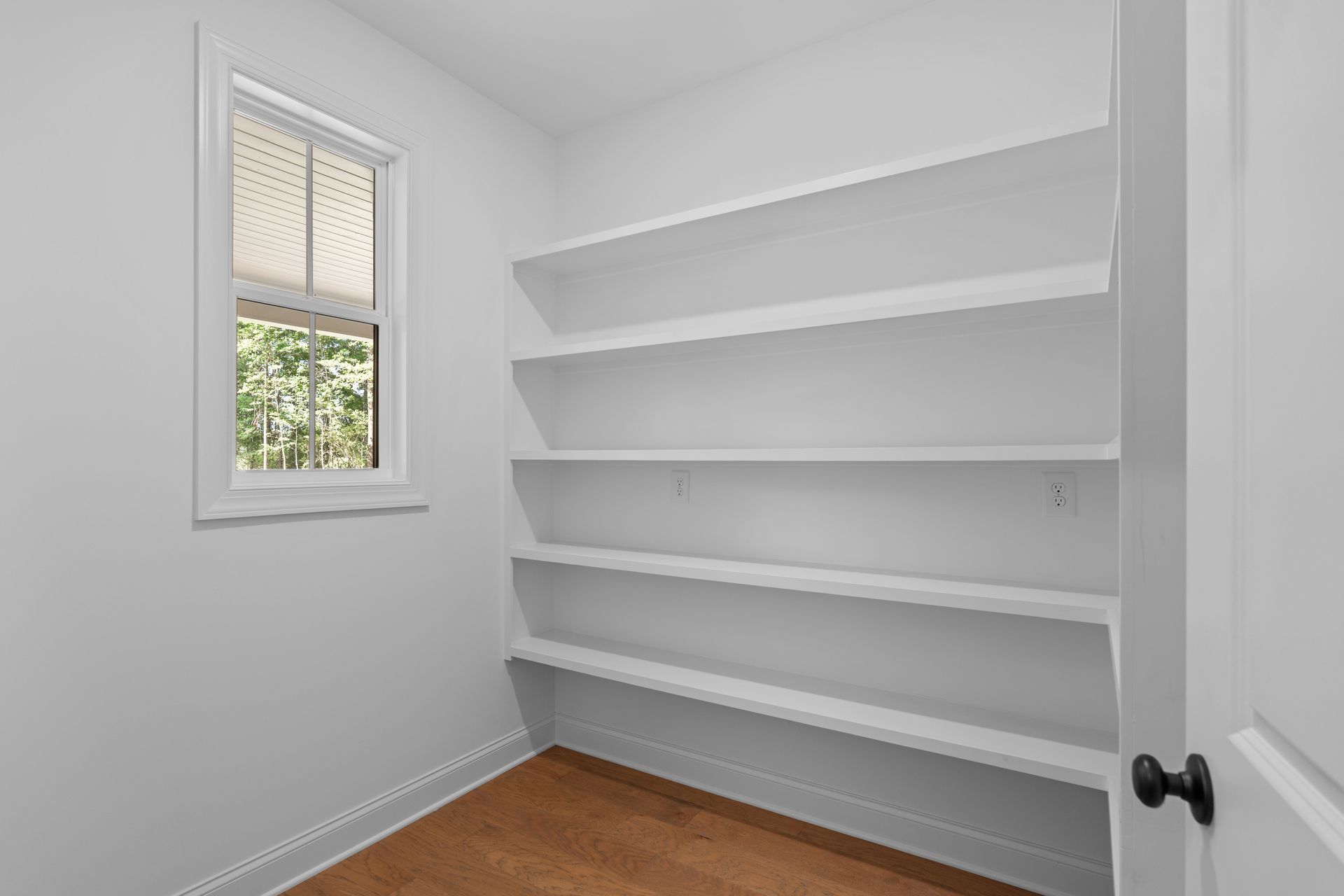 Small white closet with built-in shelves, a window, and a wooden floor.
