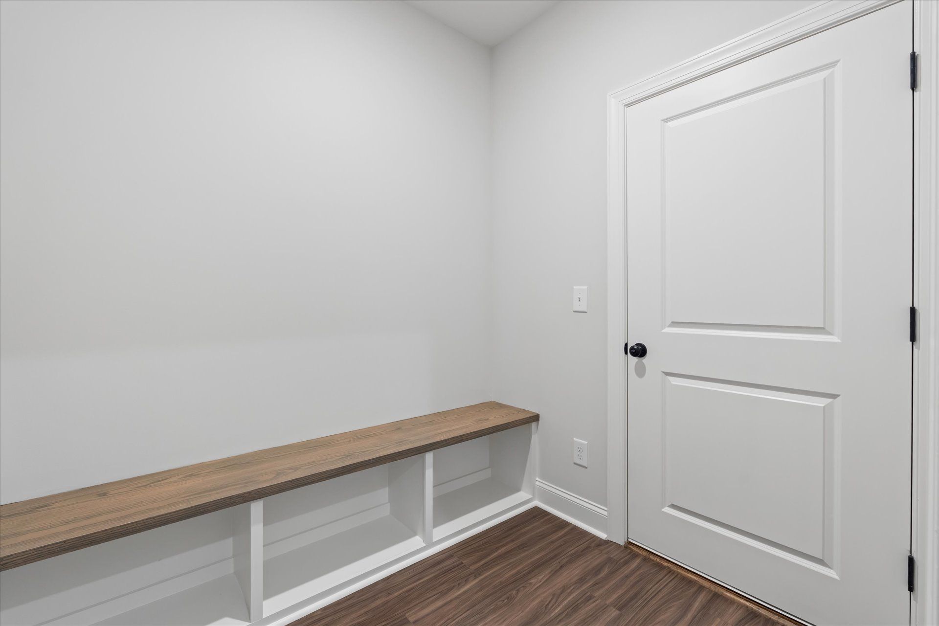 Empty white room with built-in wooden bench, white door, and wood-look floorboard floor.