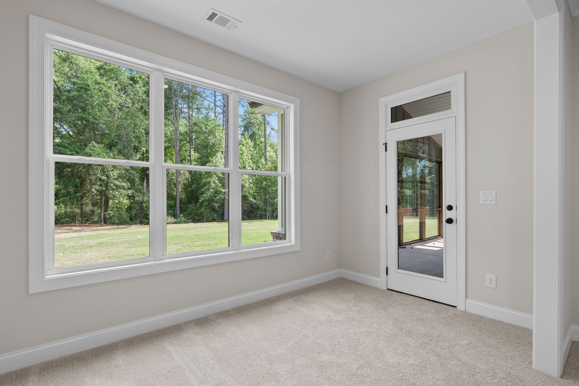 Empty beige room with large window, glass door, and carpeted floor overlooking greenery