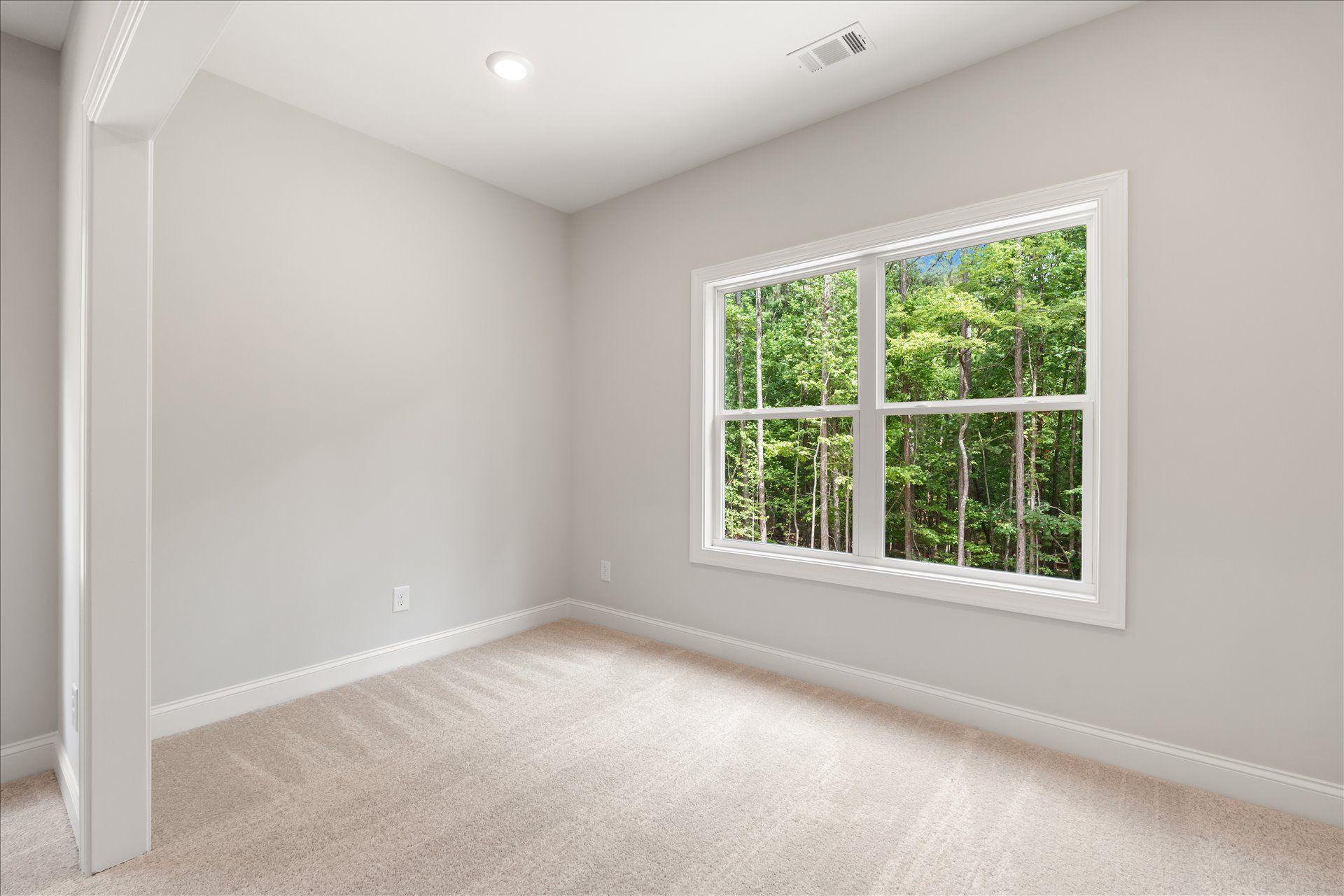 Empty beige bedroom with white trim, carpeted floor, and a large window overlooking green trees