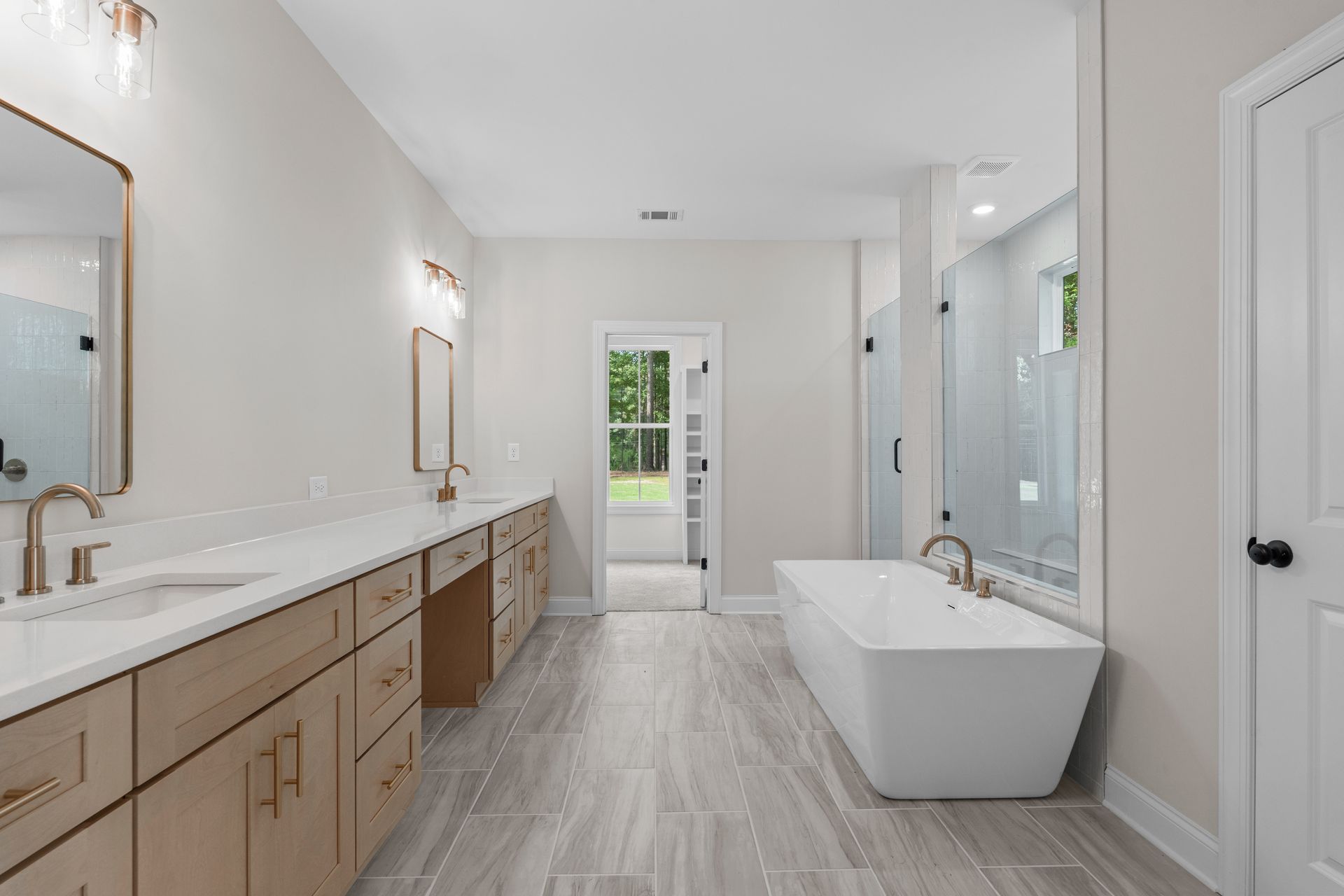 Bright modern bathroom with double vanity, freestanding tub, and light wood cabinets, viewed toward a glass door