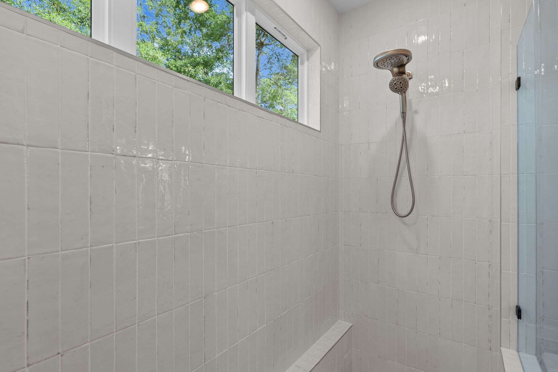 White-tiled shower with windows and a wall-mounted showerhead in a bright bathroom.