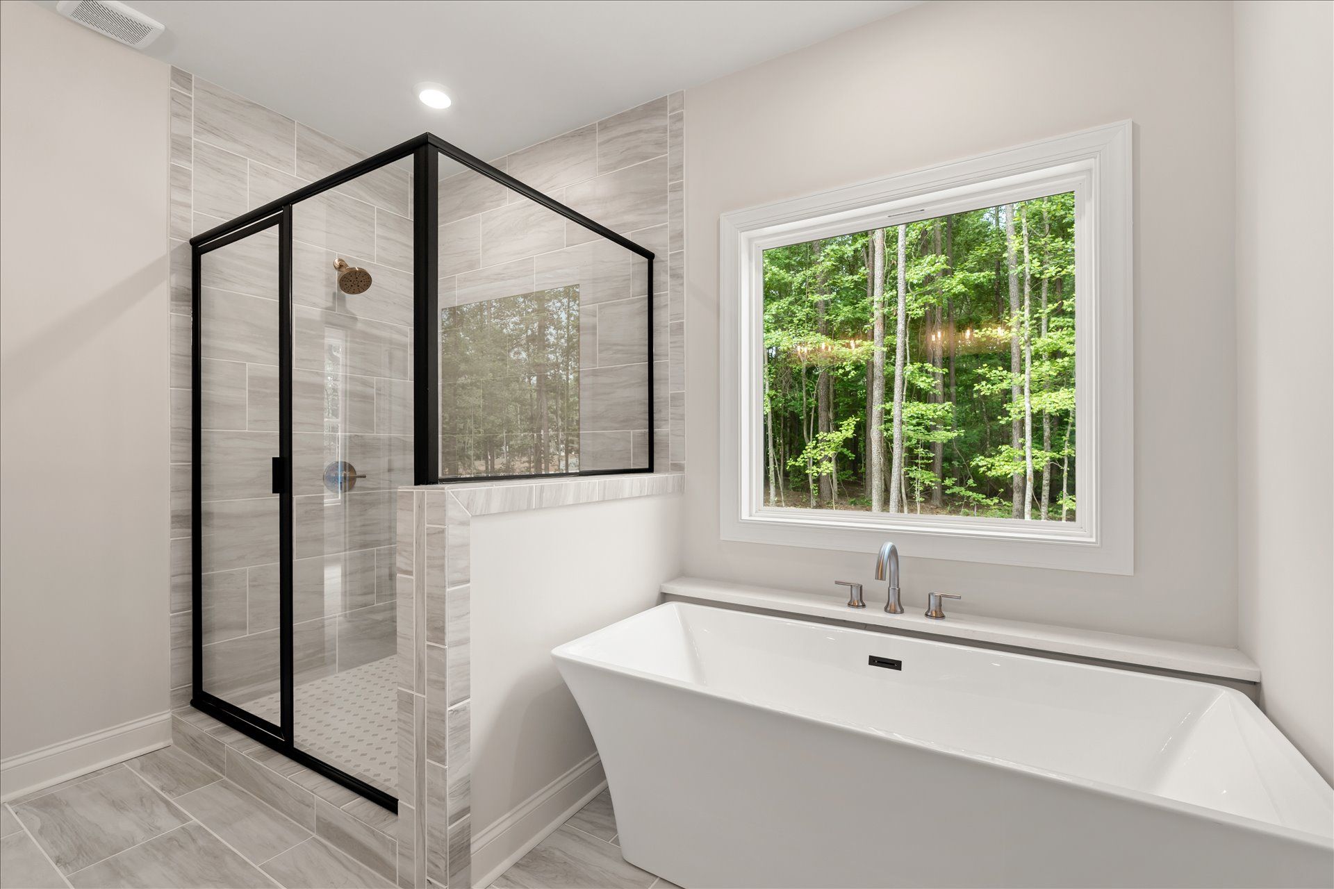 Modern bathroom with black-framed glass shower, white freestanding tub, and window overlooking greenery