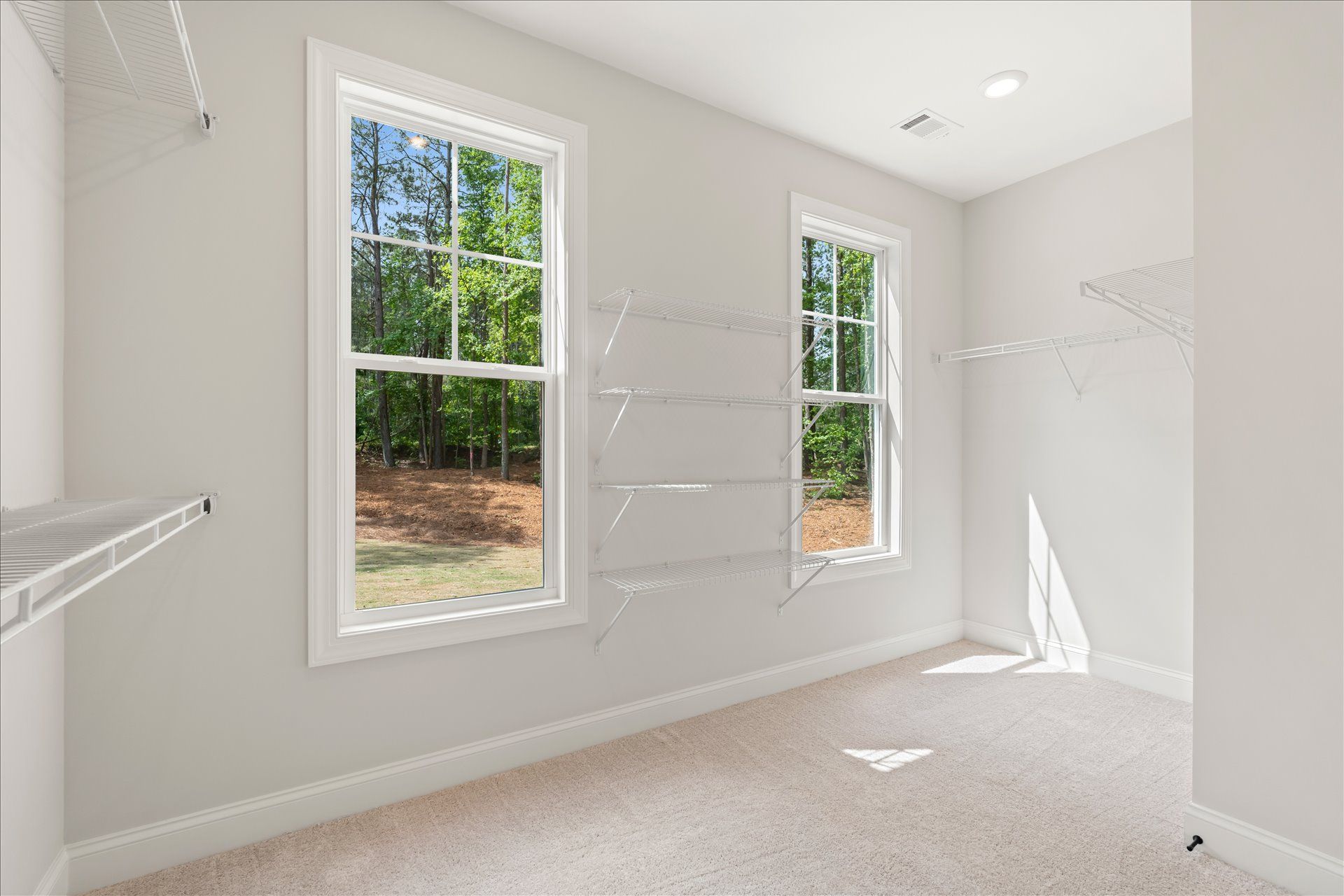 Empty bright bedroom with white walls, carpet, and two tall windows letting in daylight.