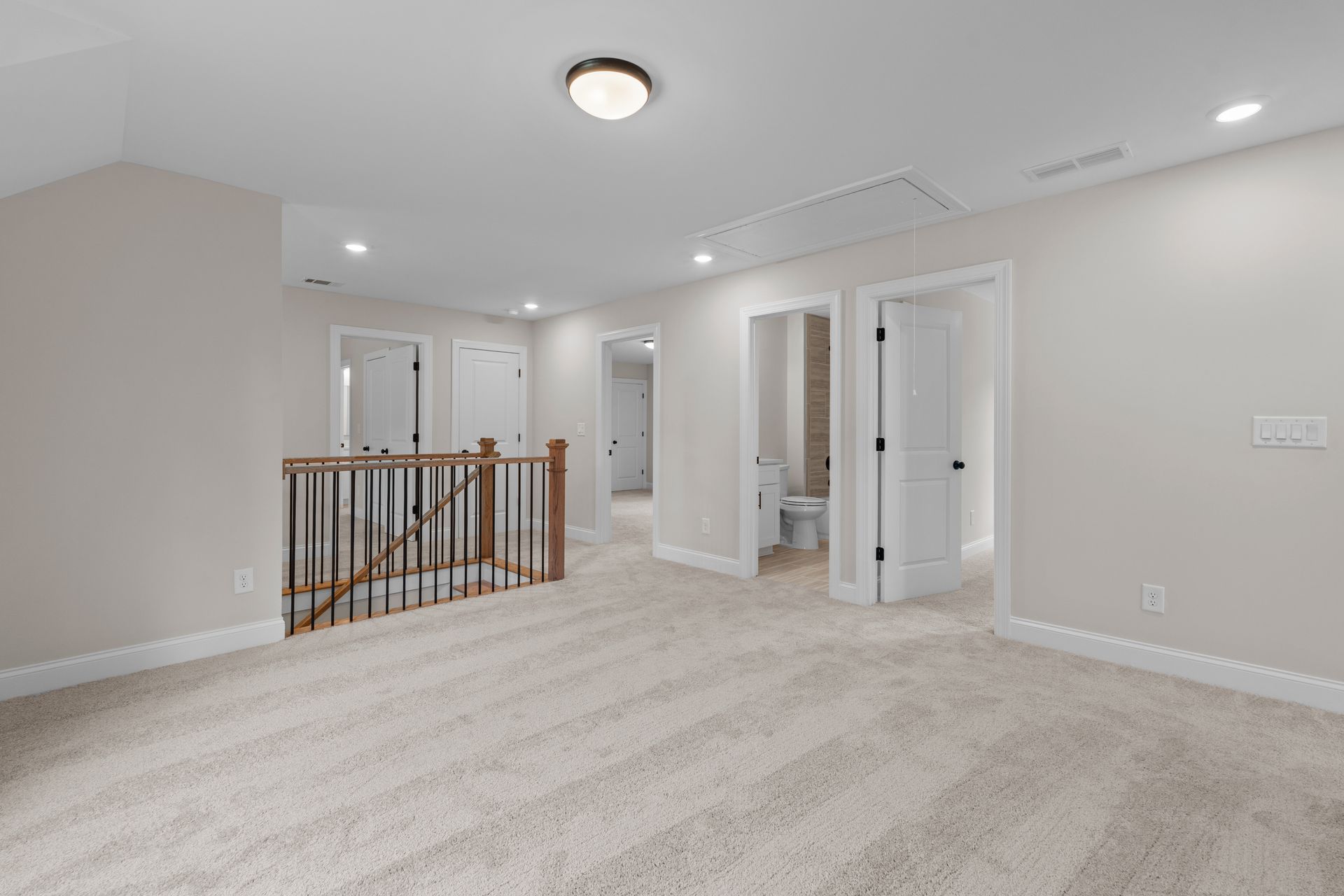 Empty beige carpeted hallway and landing with white walls, recessed lights, and wooden stair railing