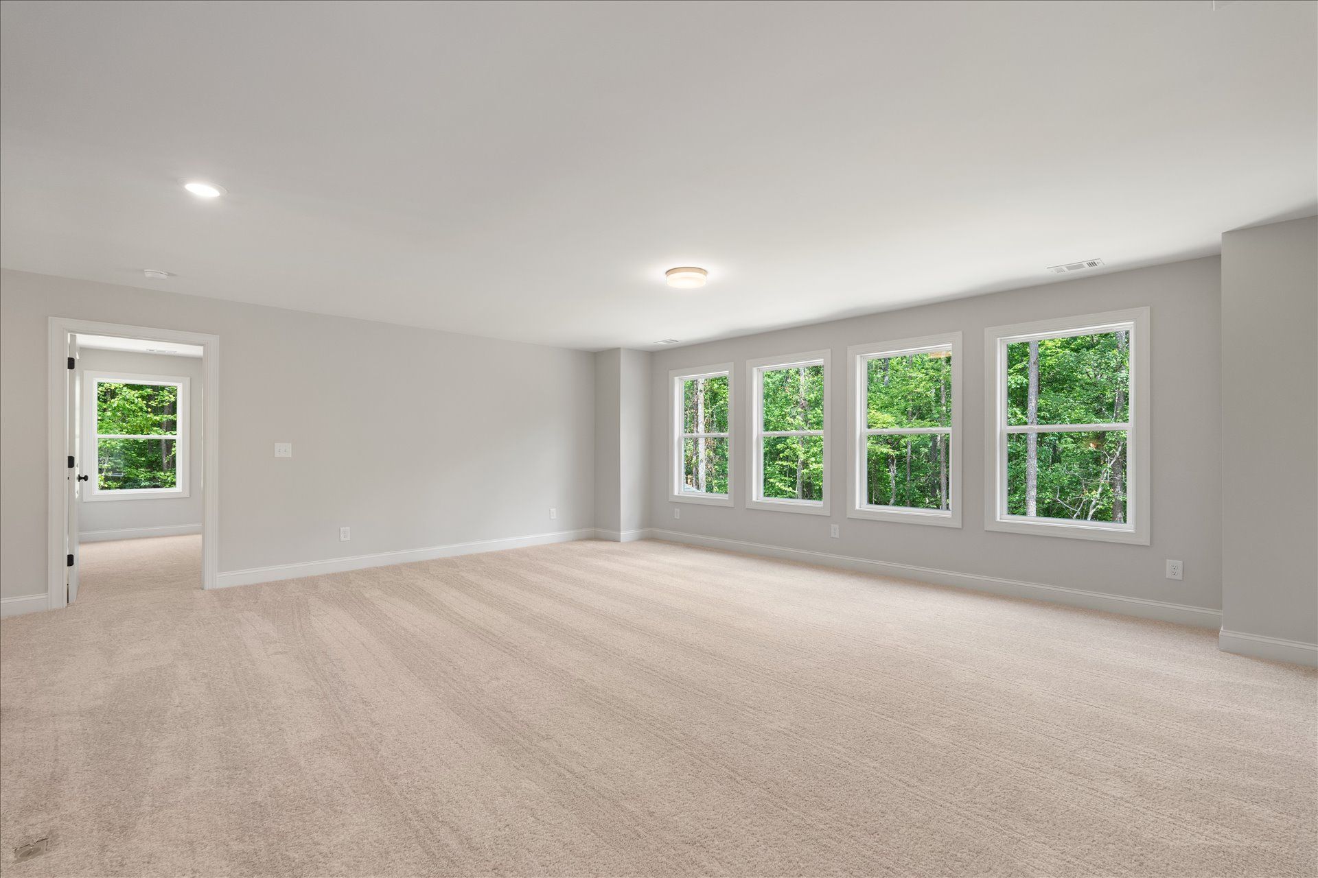 Bright empty living room with beige carpet, white walls, and large windows overlooking green trees