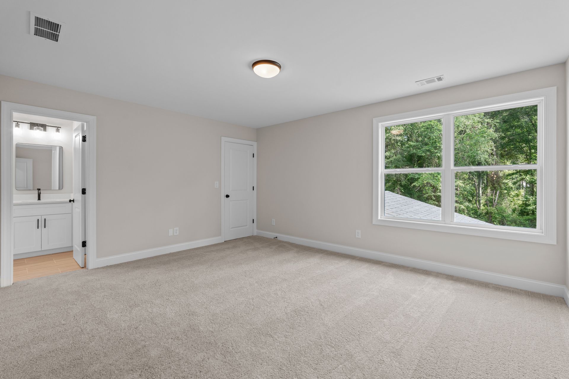 Empty beige bedroom with carpet, white walls, closet door, and a large window overlooking greenery
