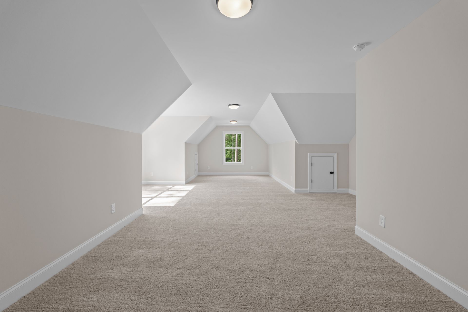 Empty carpeted attic room with white walls, sloped ceiling, and a small window