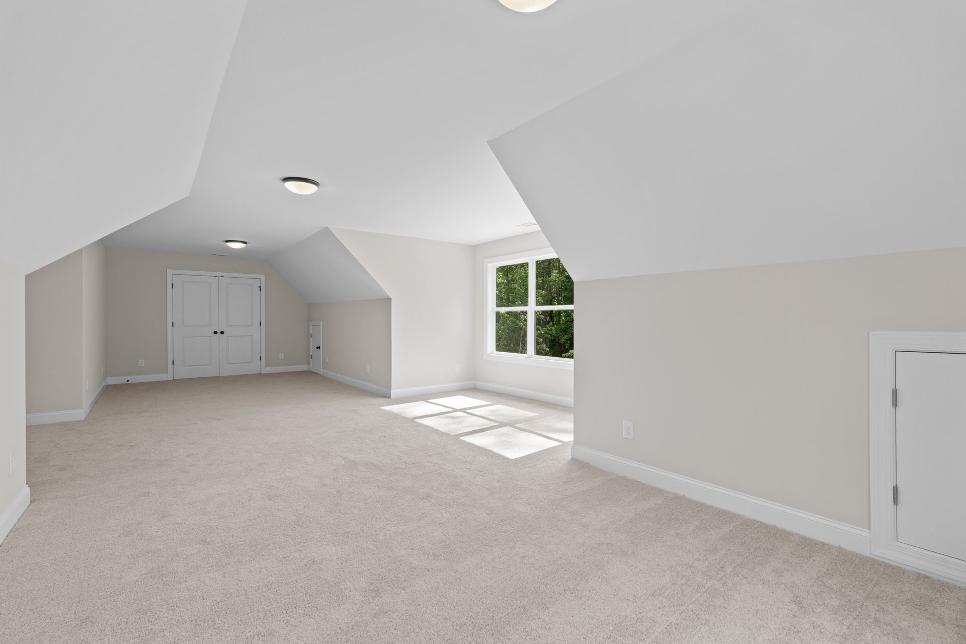 Bright empty attic room with white walls, beige carpet, and a large window letting in sunlight.