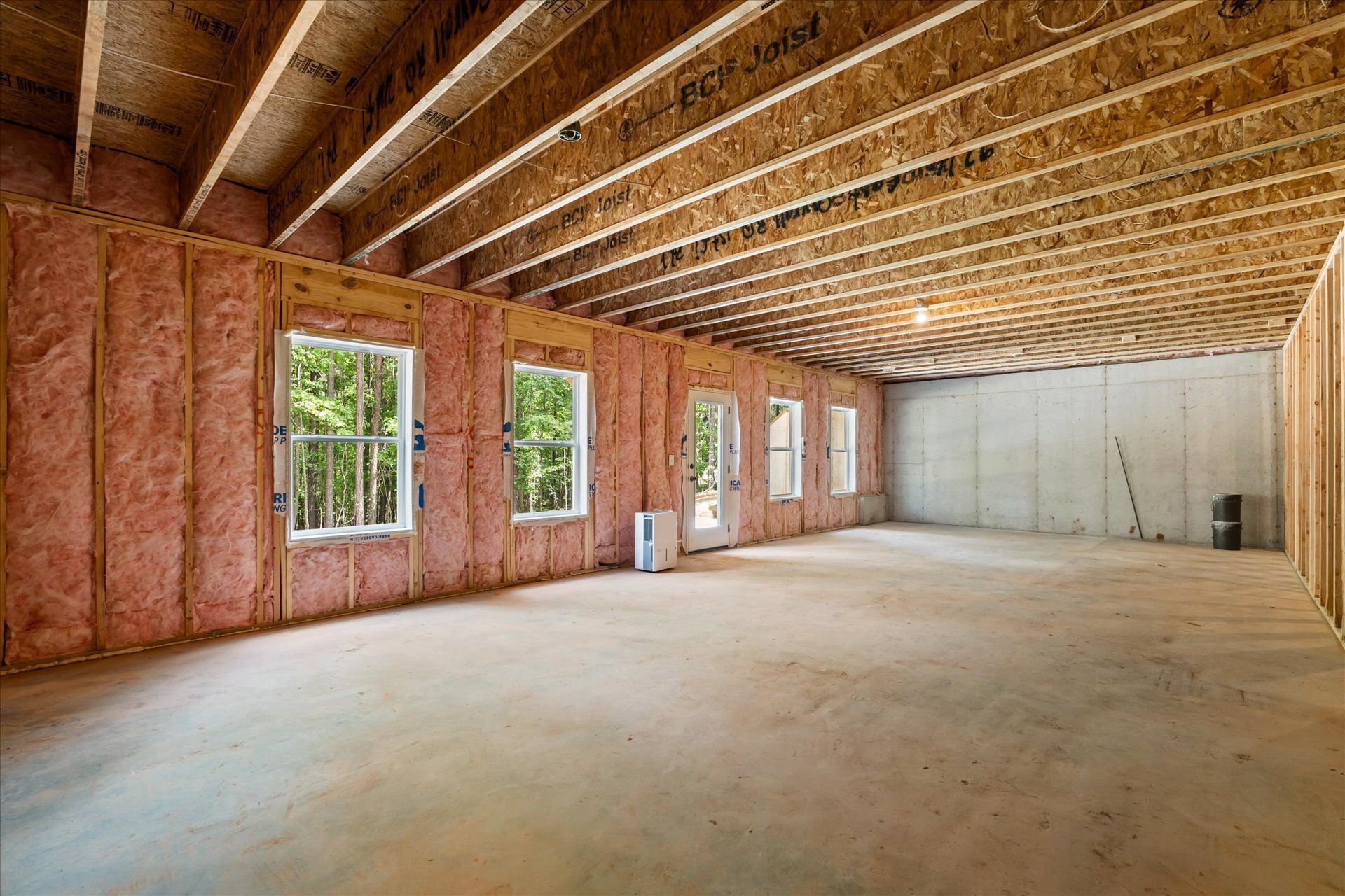 Unfinished interior with exposed wood framing, pink insulation, concrete floor, and windows along one wall