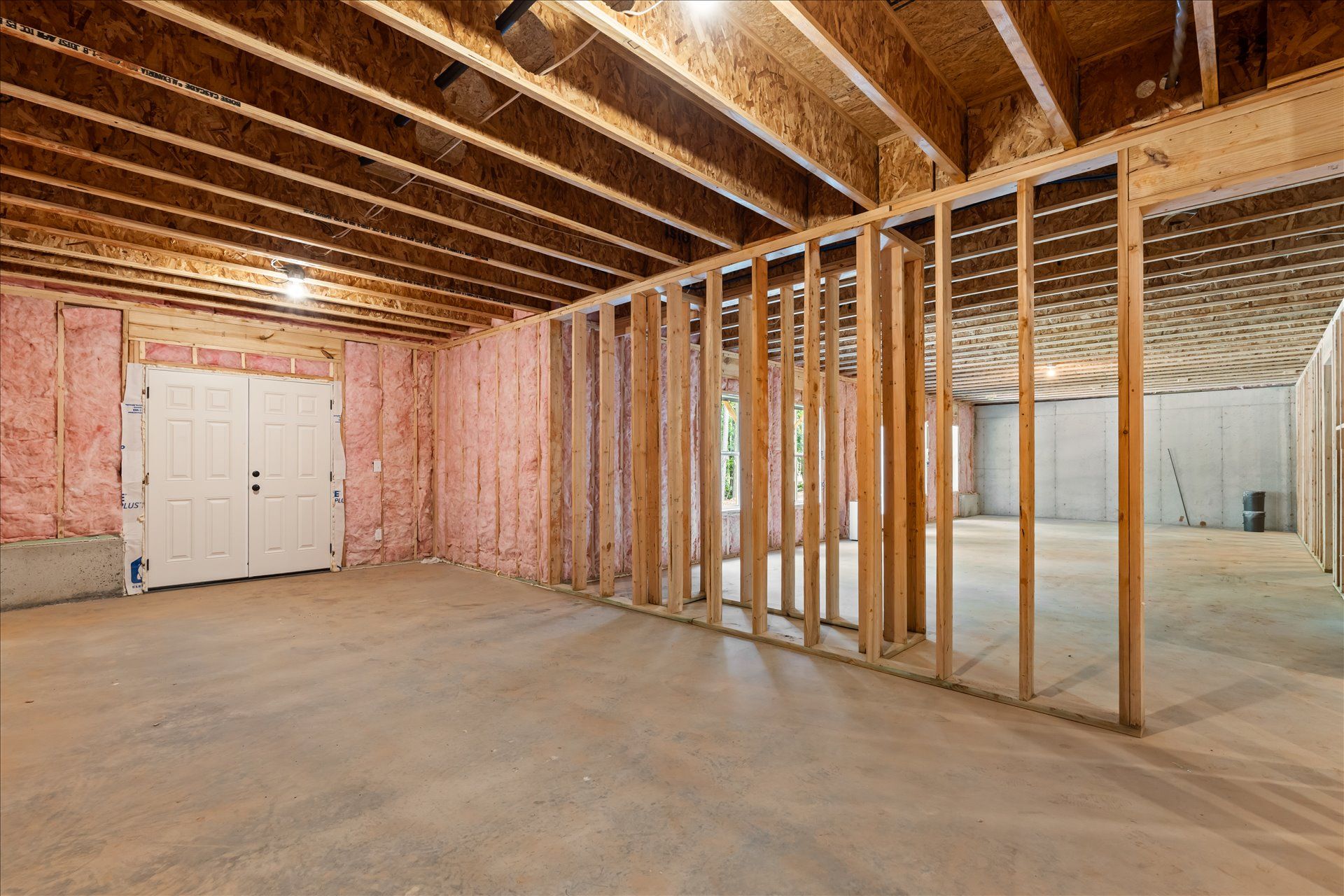 Unfinished basement with exposed studs, insulation, and ceiling beams, featuring a white door and concrete floor
