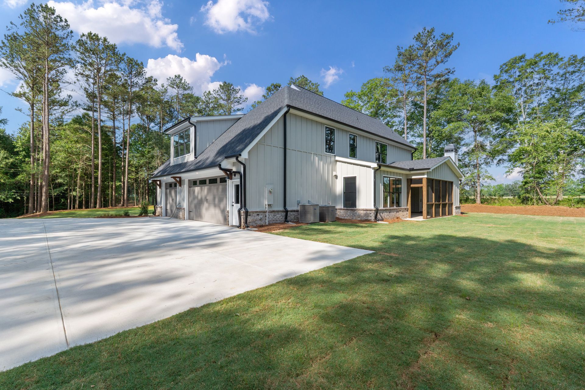 Modern gray house with a large concrete driveway, green lawn, and trees under a blue sky