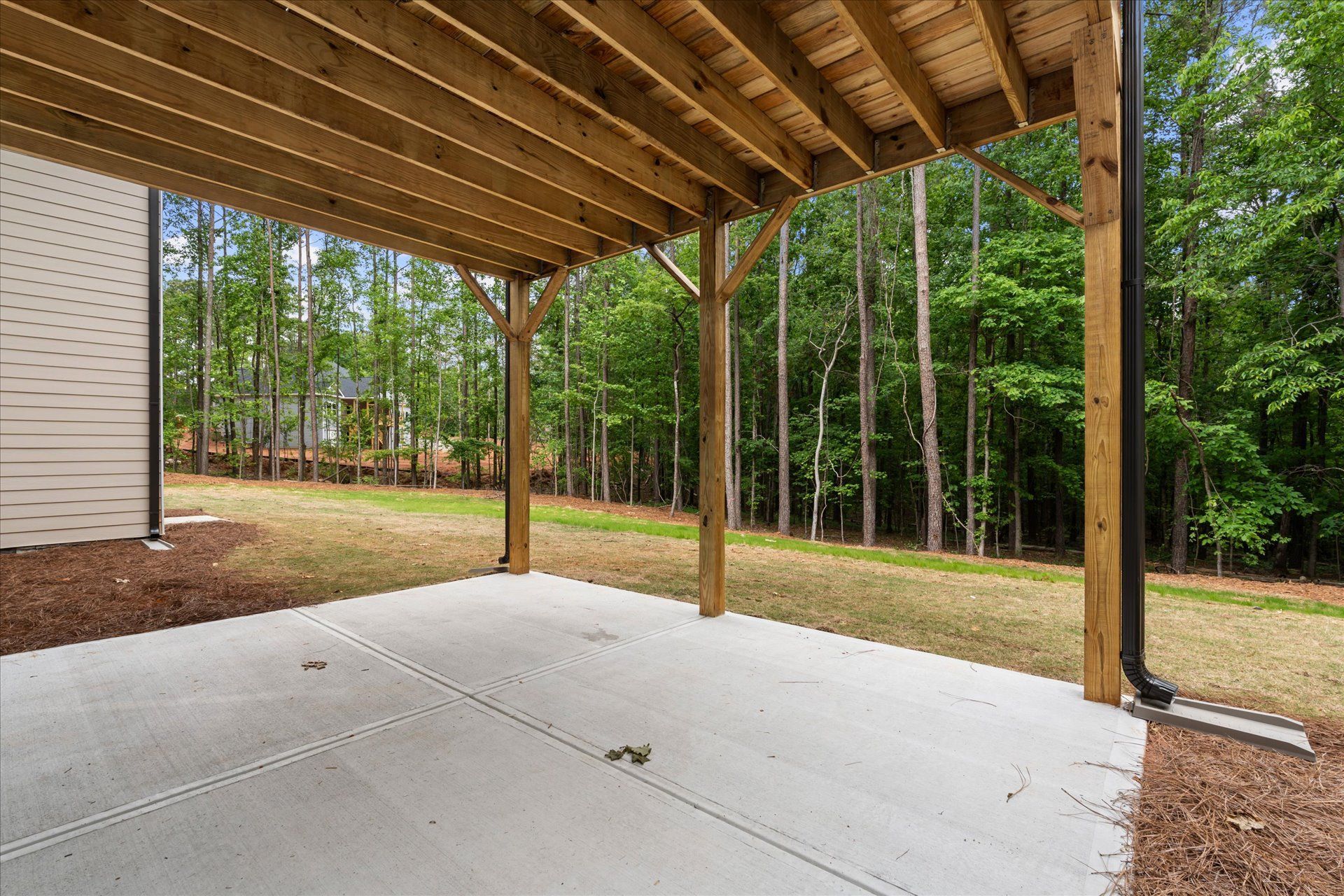 Covered backyard patio with concrete slab, wood posts, and a wooded yard beyond