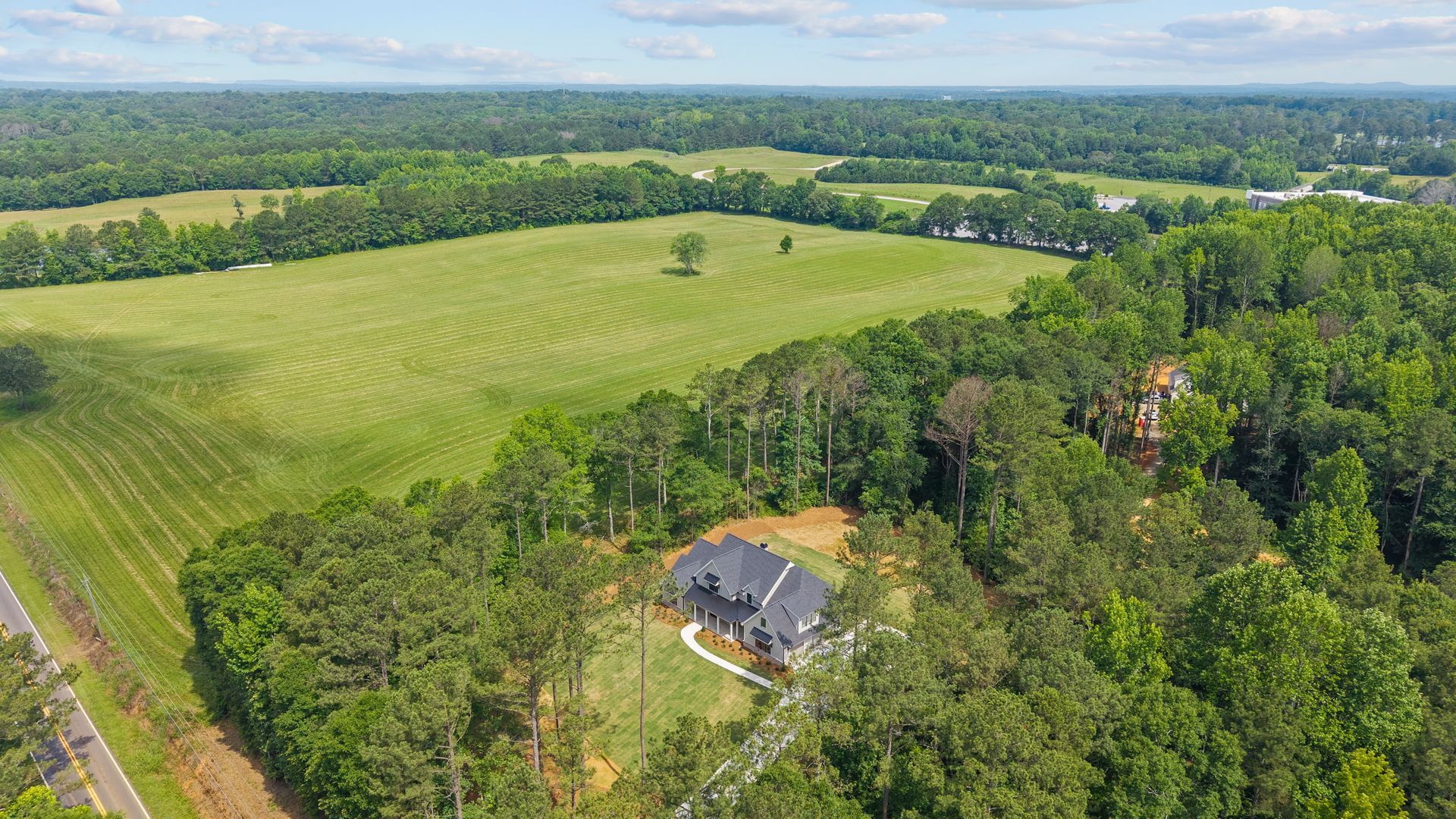 Aerial view of a house beside dense trees and a large green field under a partly cloudy sky.