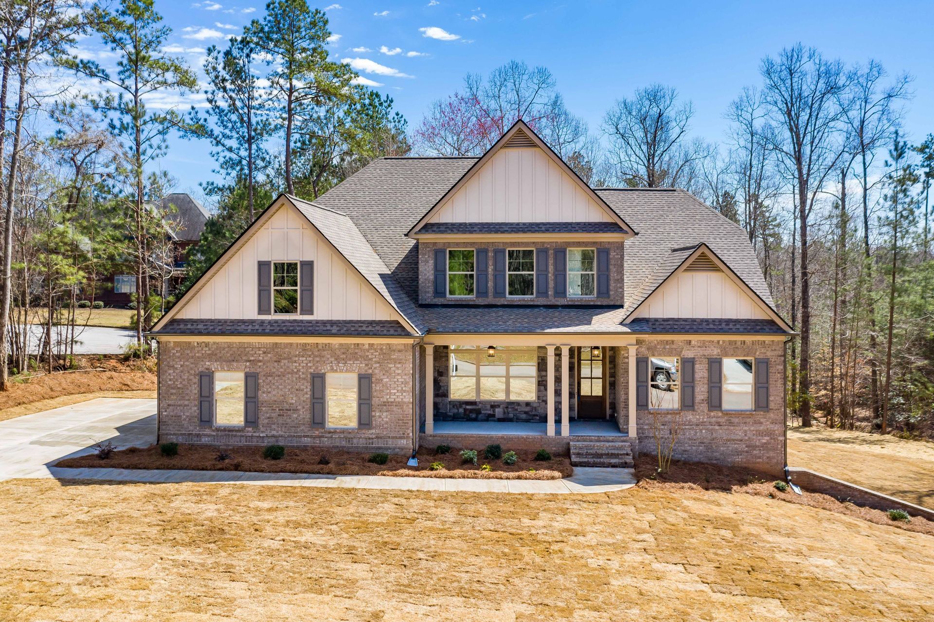 Two-story brick house with gabled roof and front porch in a wooded lot under a clear blue sky