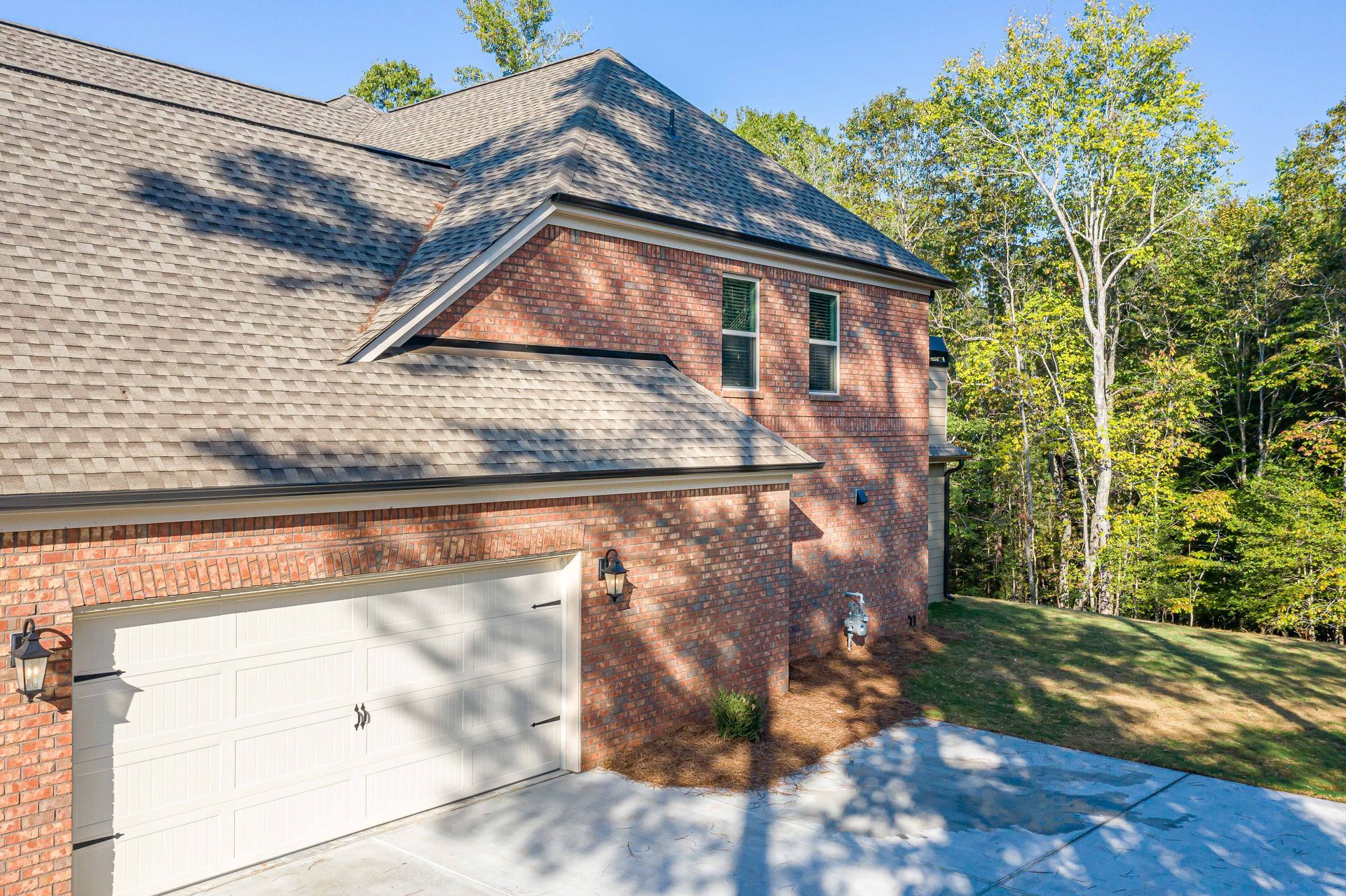 Brick house with white garage door, sloped roof, and trees in the yard on a sunny day