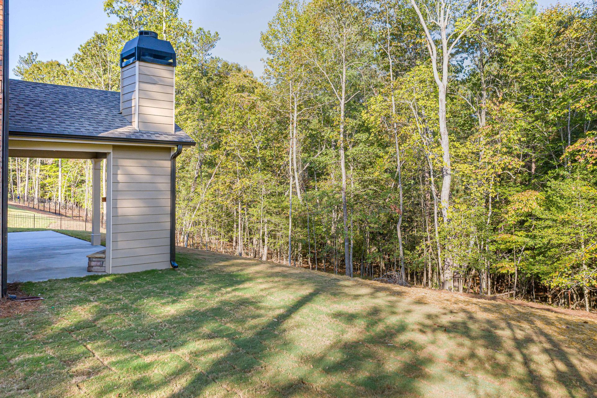 Backyard lawn beside a beige house with a chimney, bordered by tall trees and woods