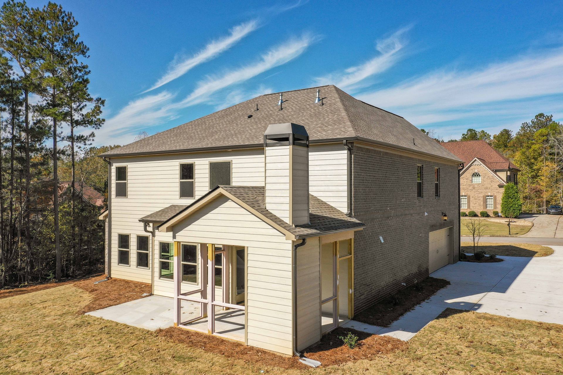 Two-story beige house with brown roof, front porch, and driveway in a wooded neighborhood.