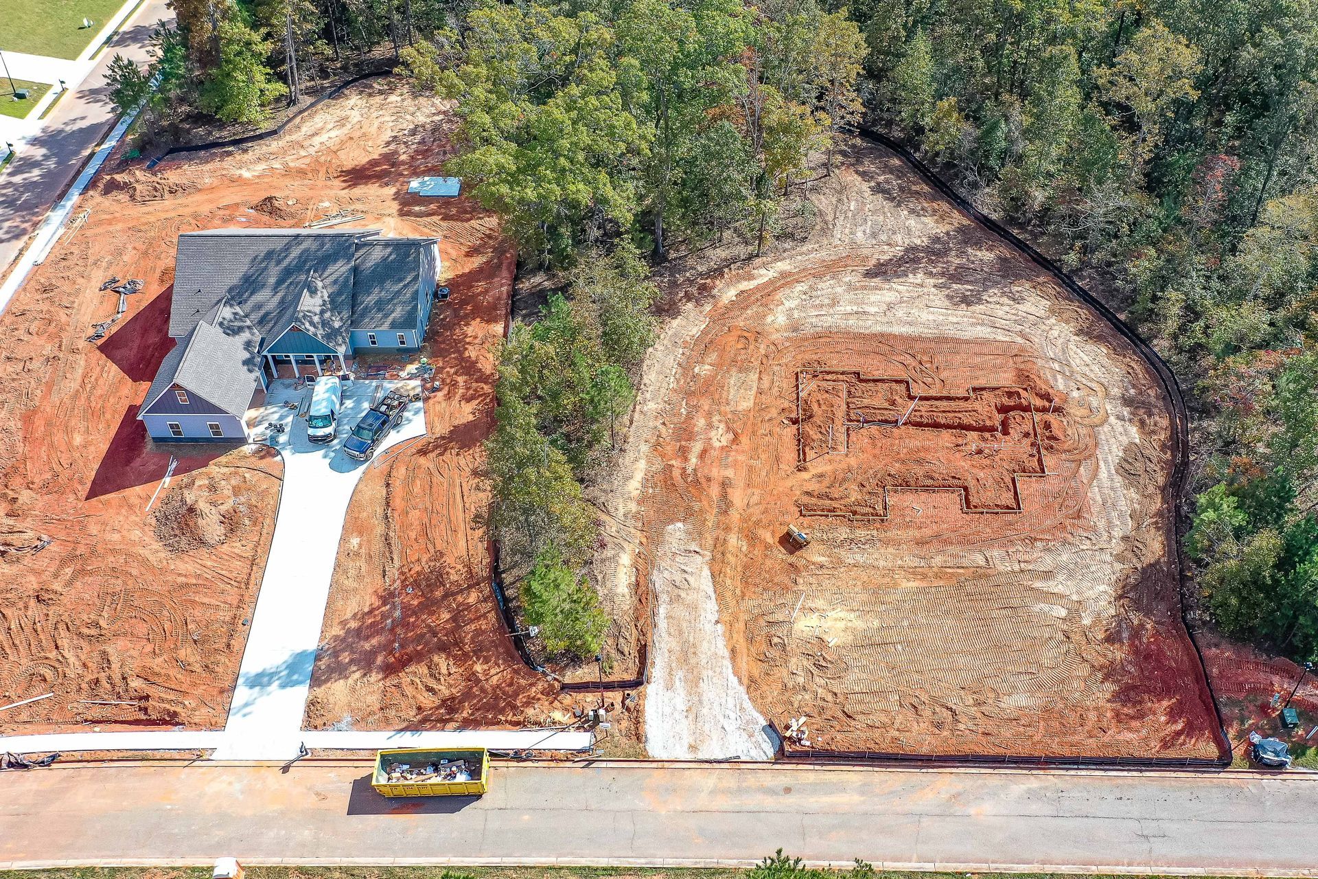 Aerial view of a house under construction beside a cleared dirt lot and road, surrounded by trees.