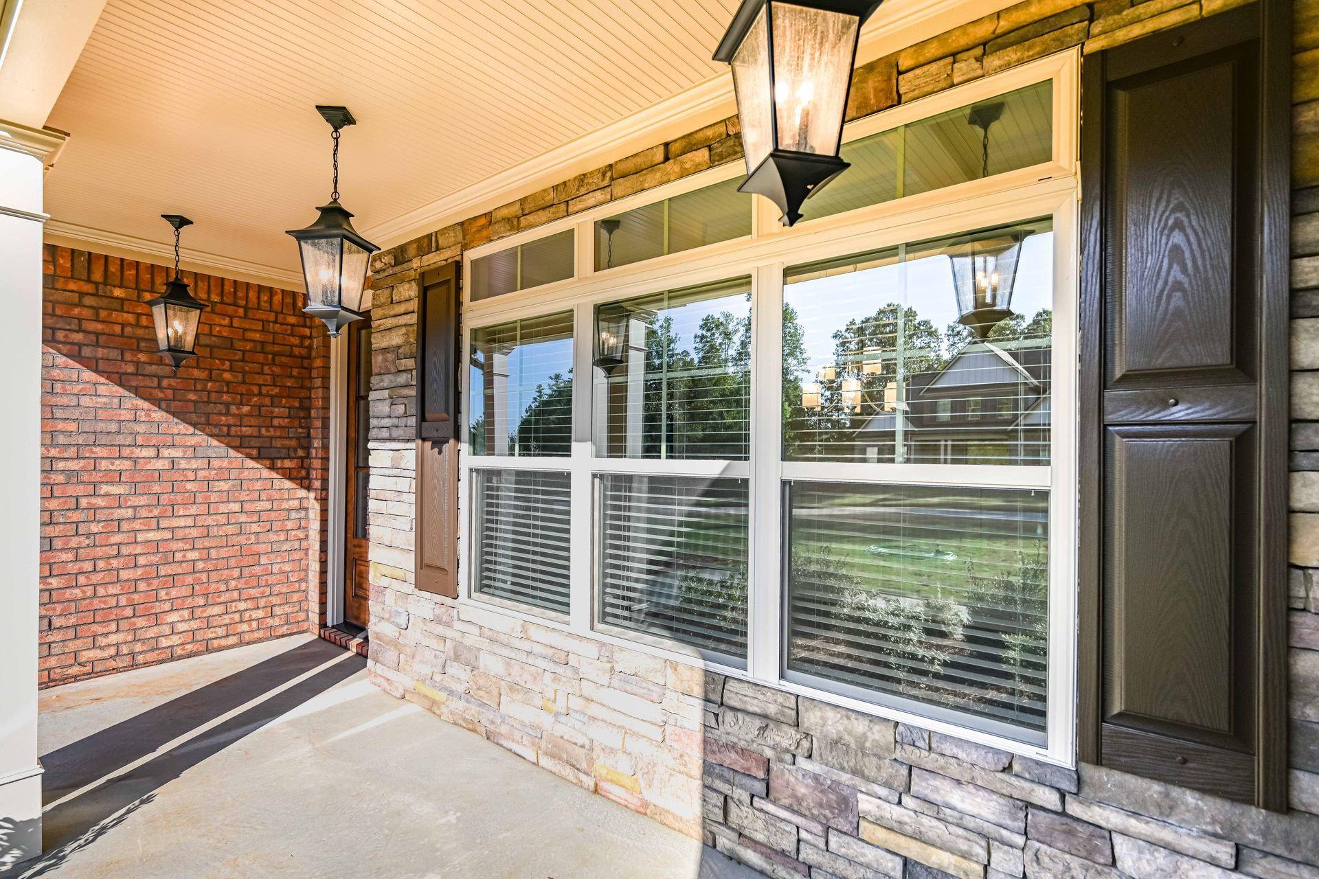 Covered front porch with brick wall, hanging lanterns, and large windows beside a grassy yard.