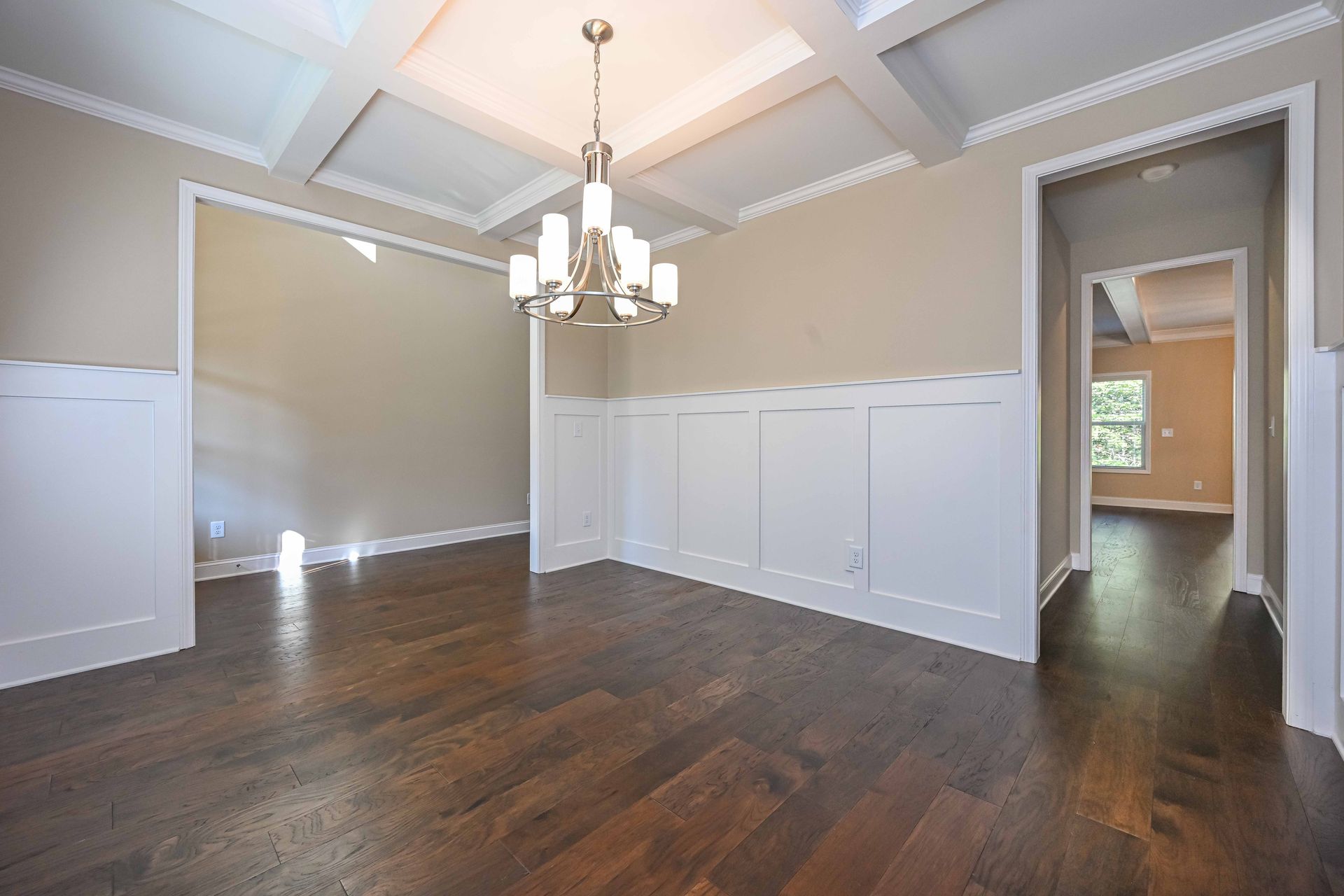 Empty dining room with dark hardwood floors, beige walls, white wainscoting, and a chandelier under a tray ceiling