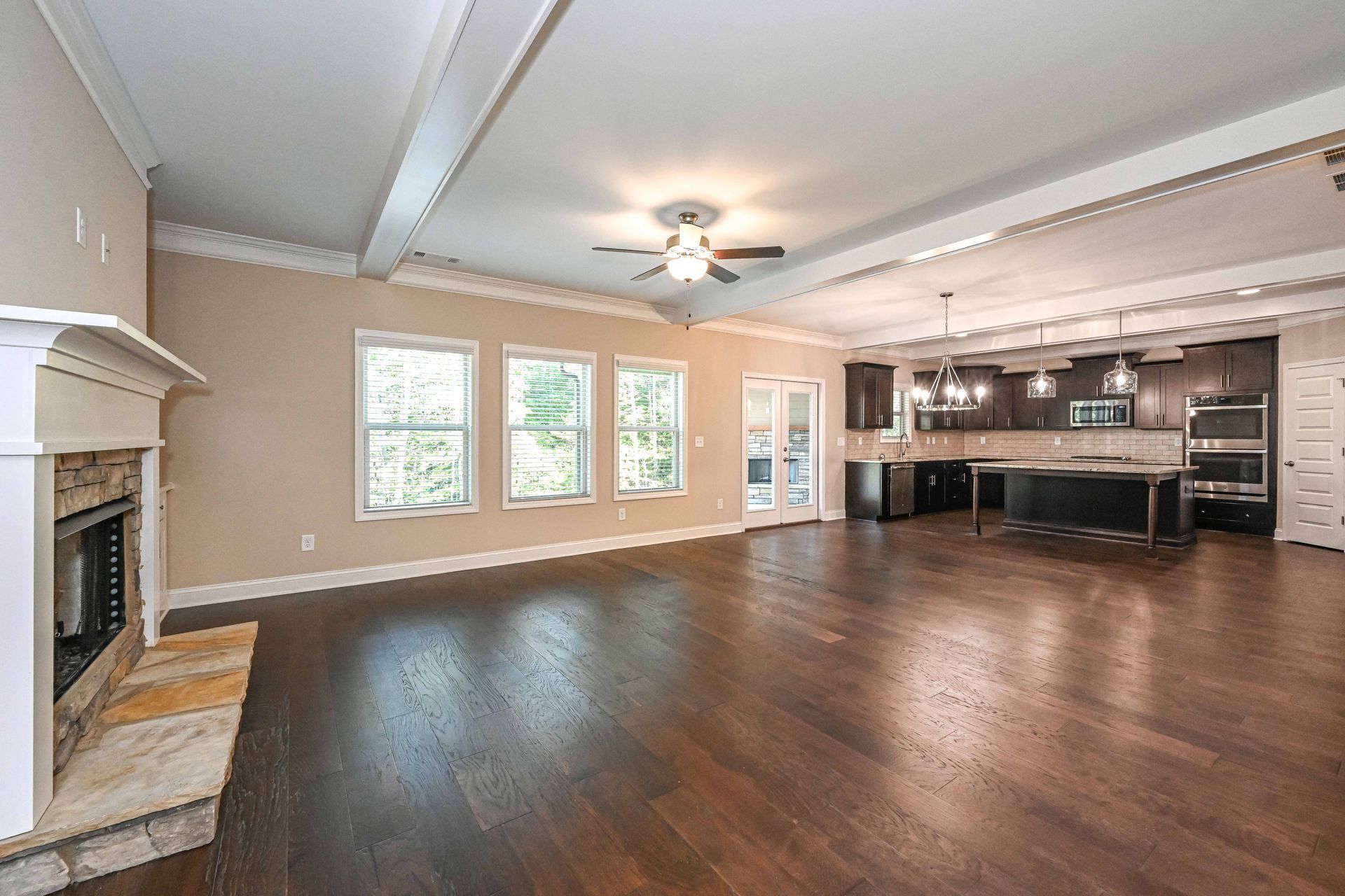 Spacious empty living room with dark wood floors, fireplace, ceiling fan, and open kitchen area
