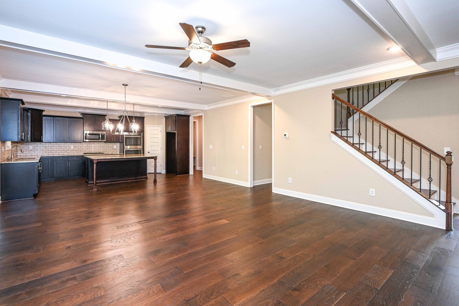 Empty living room with dark wood floors, open kitchen, ceiling fan, and staircase on the right