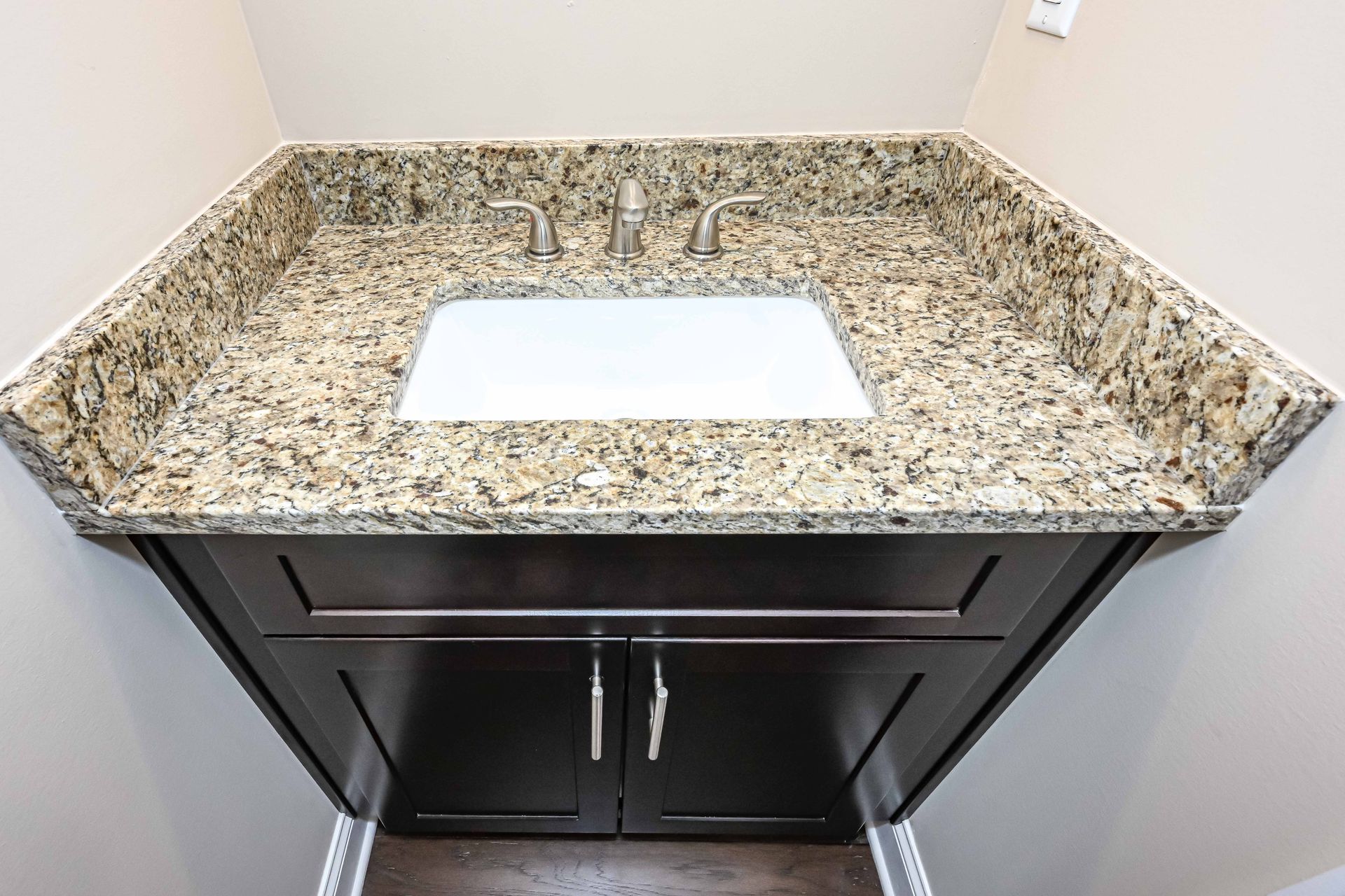 Bathroom vanity with granite countertop, white sink, and dark cabinets against beige walls