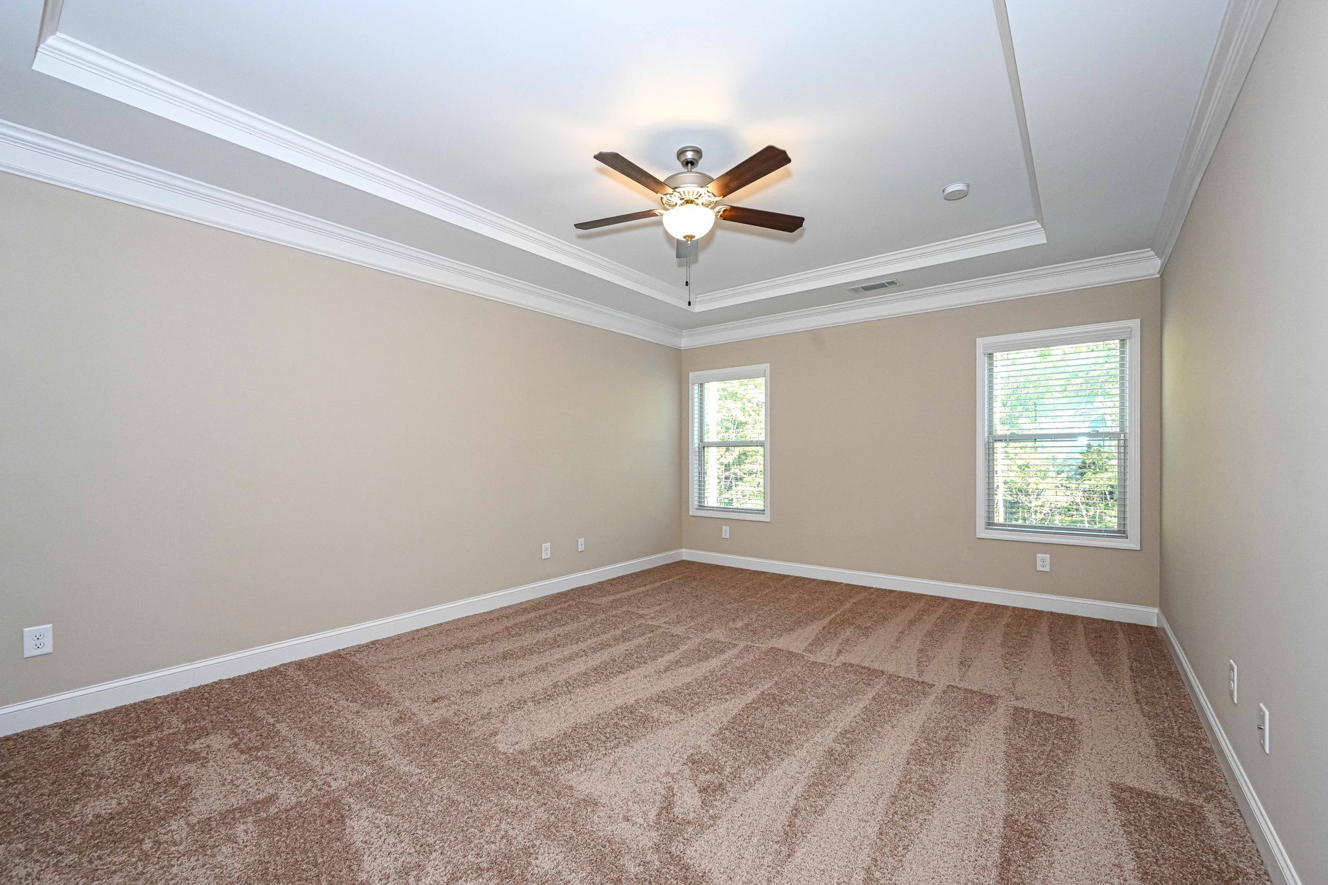 Empty beige bedroom with carpet, ceiling fan, and two windows with white trim.