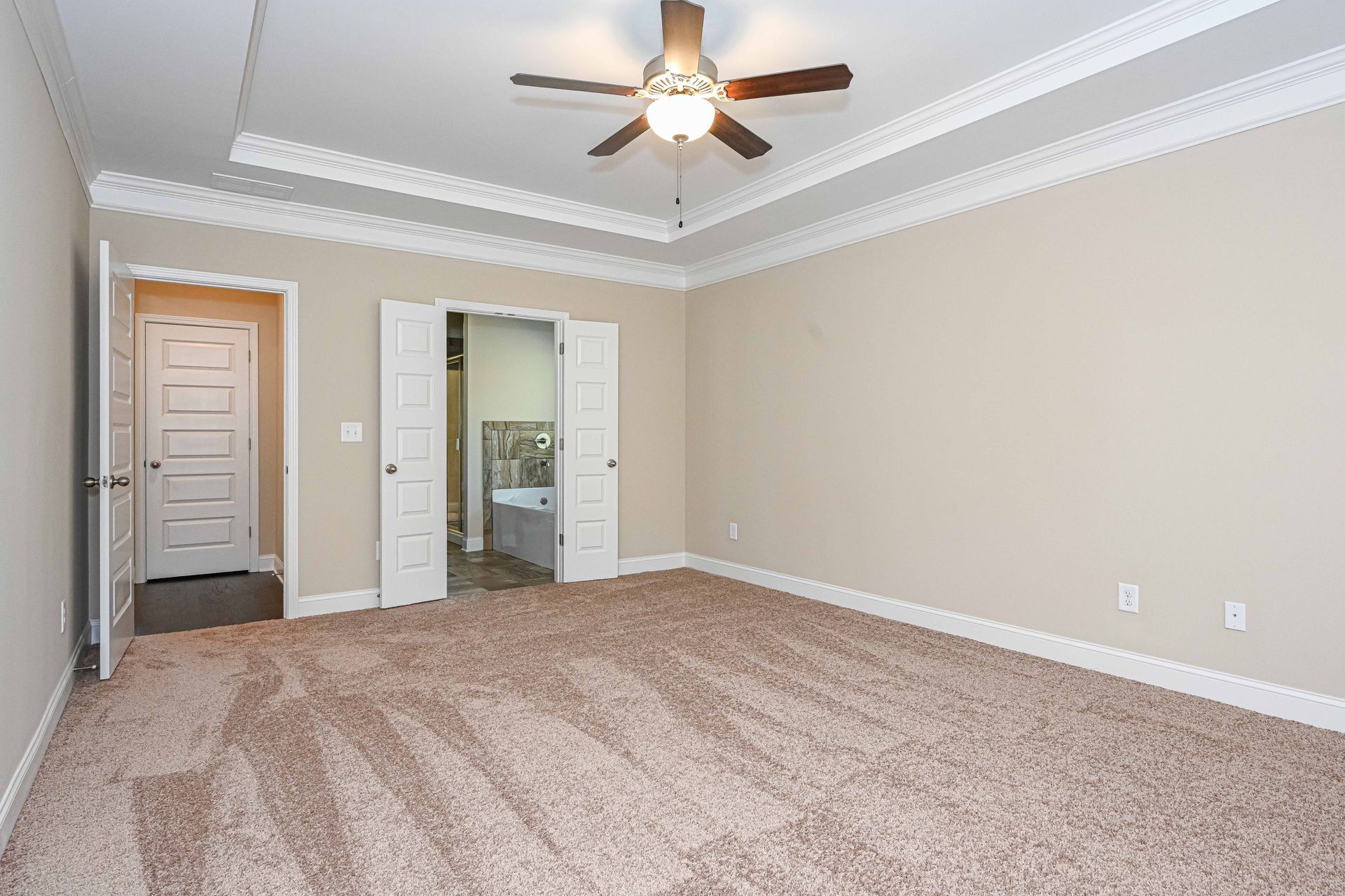 Empty beige bedroom with carpet, ceiling fan, crown molding, and open doorway to a bathroom.