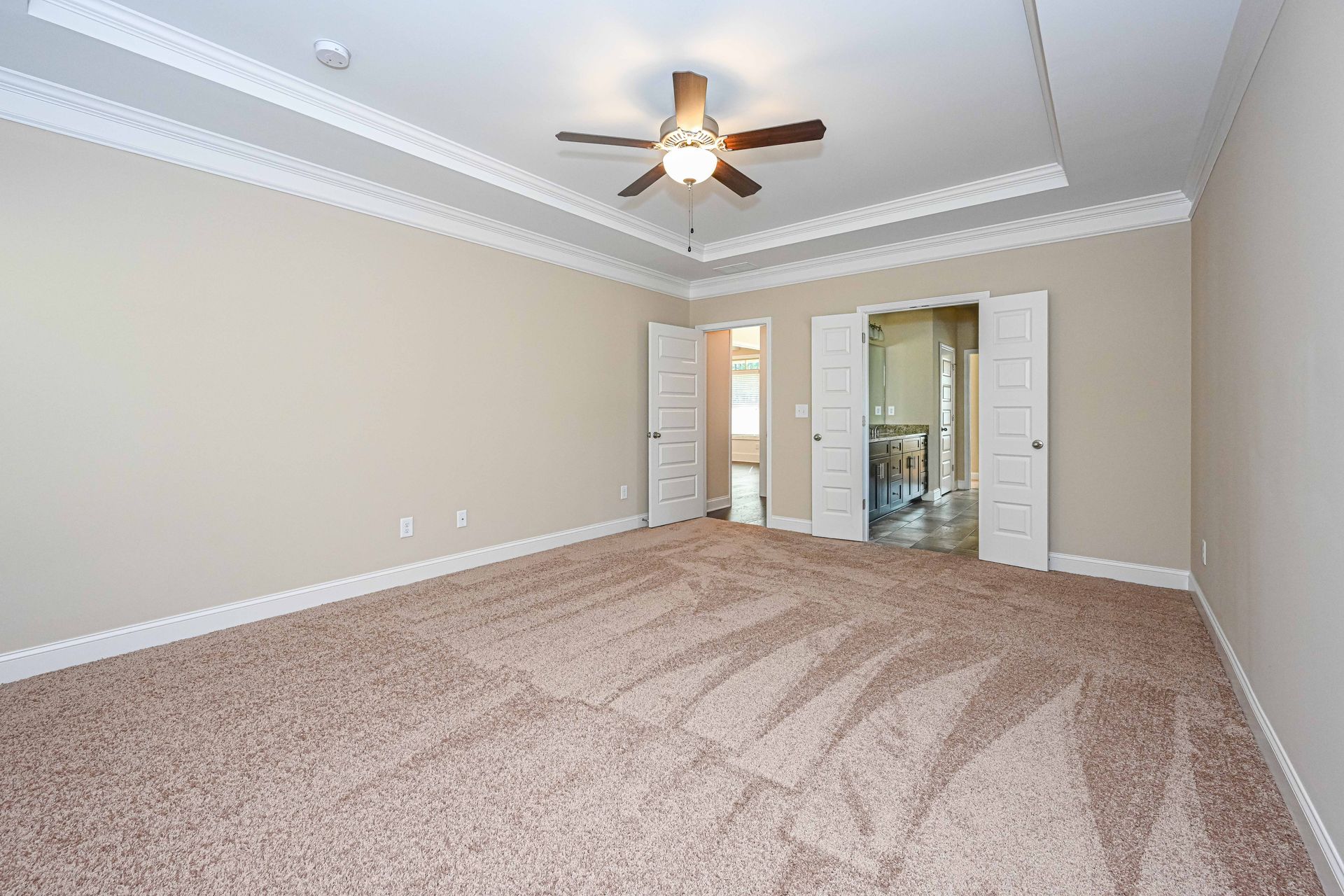 Empty beige carpeted bedroom with tray ceiling, ceiling fan, and open doorway to bathroom