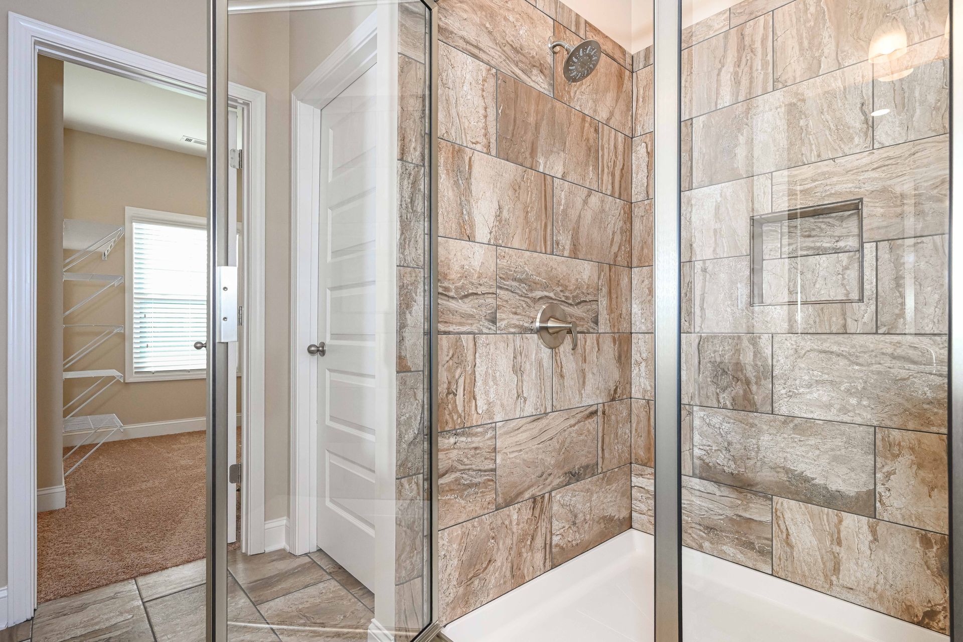 Bathroom with glass shower doors, beige stone tile walls, and a mirrored closet door reflected in the hallway