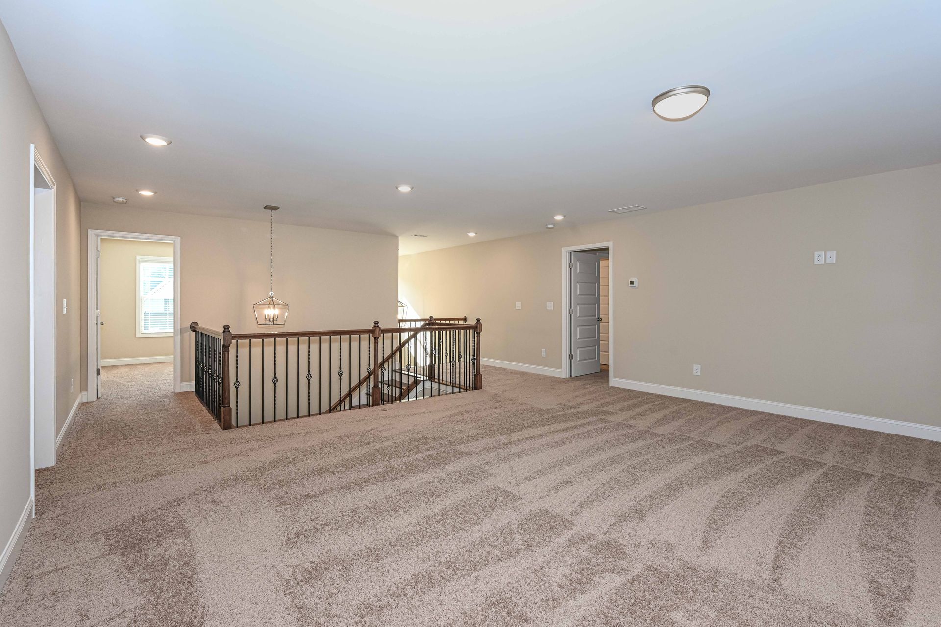 Spacious beige carpeted living room with staircase railing, recessed lights, and doorway to hall