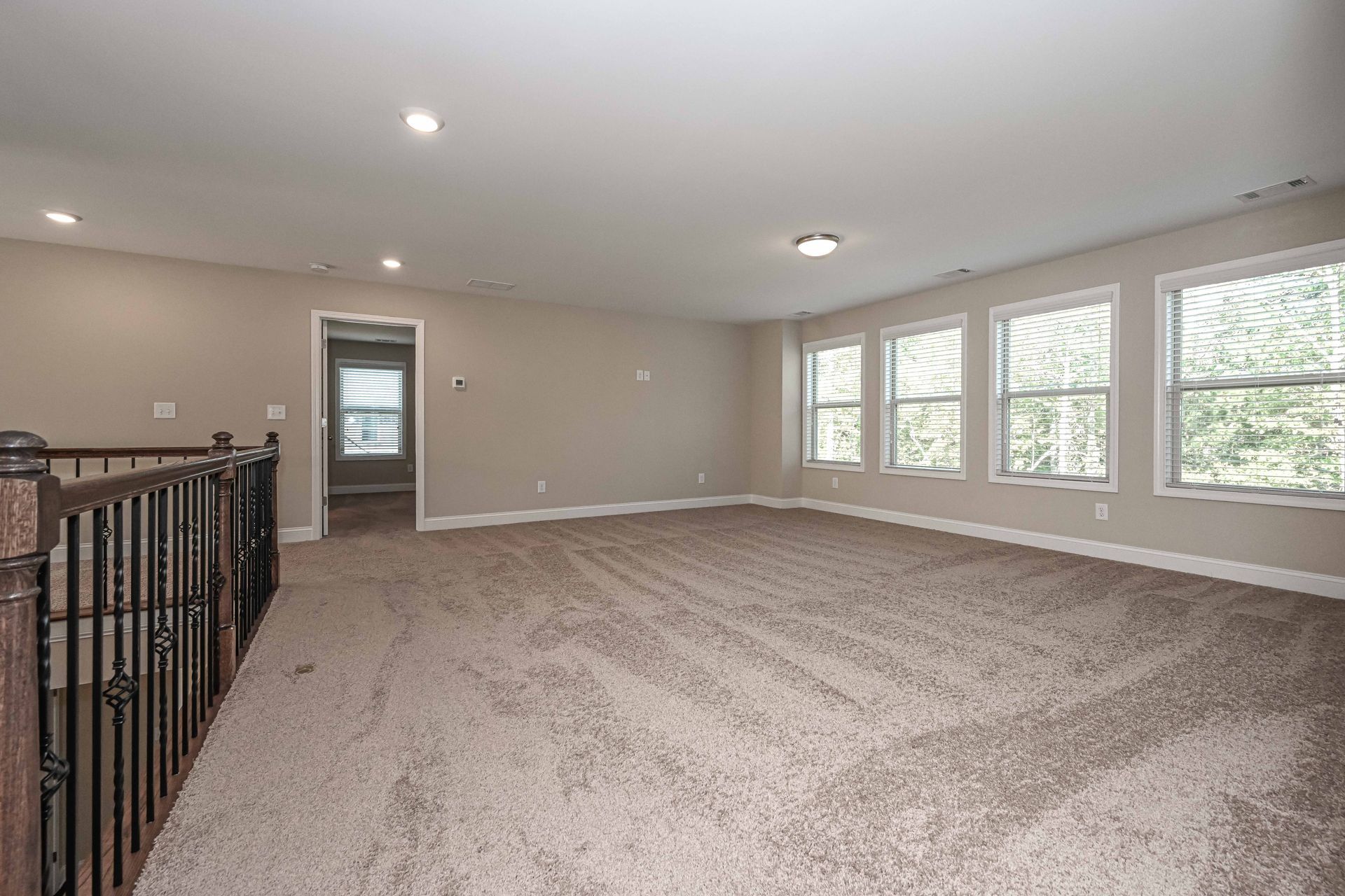 Spacious empty carpeted room with beige walls, white trim, windows, and a staircase railing on the left