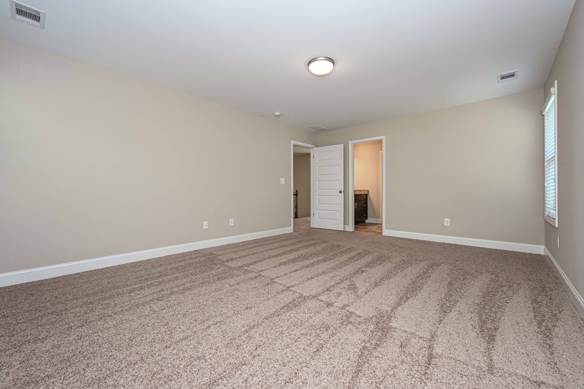 Empty beige carpeted bedroom with white walls, ceiling light, and open doorway to hallway