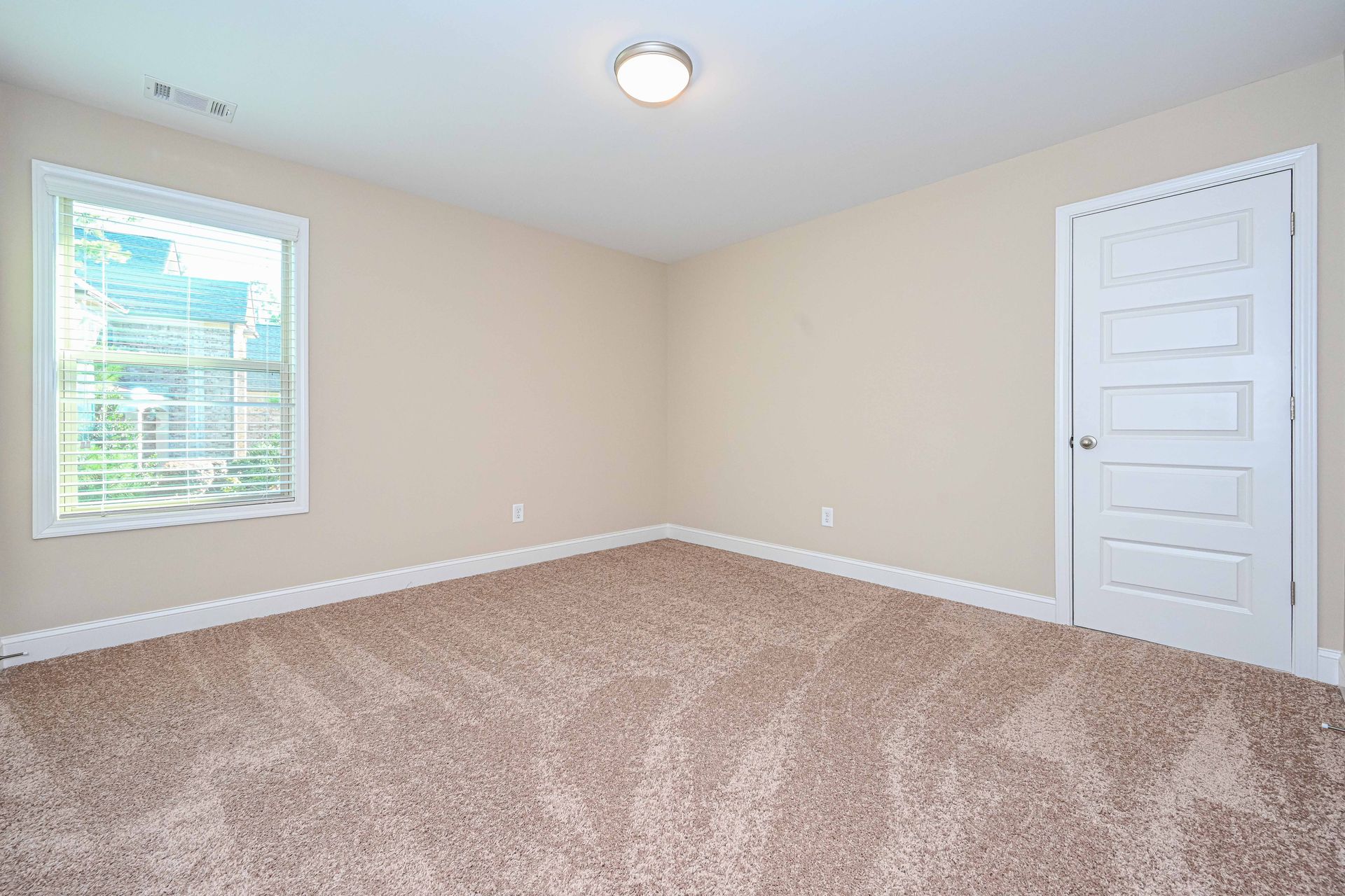 Empty beige bedroom with carpet, window, ceiling light, and white door